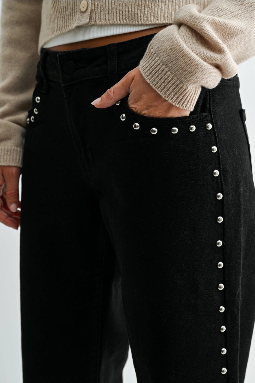 woman wearing black jeans with silver studs and a cream sweater in front of a white background with her hand in the front pocket. 