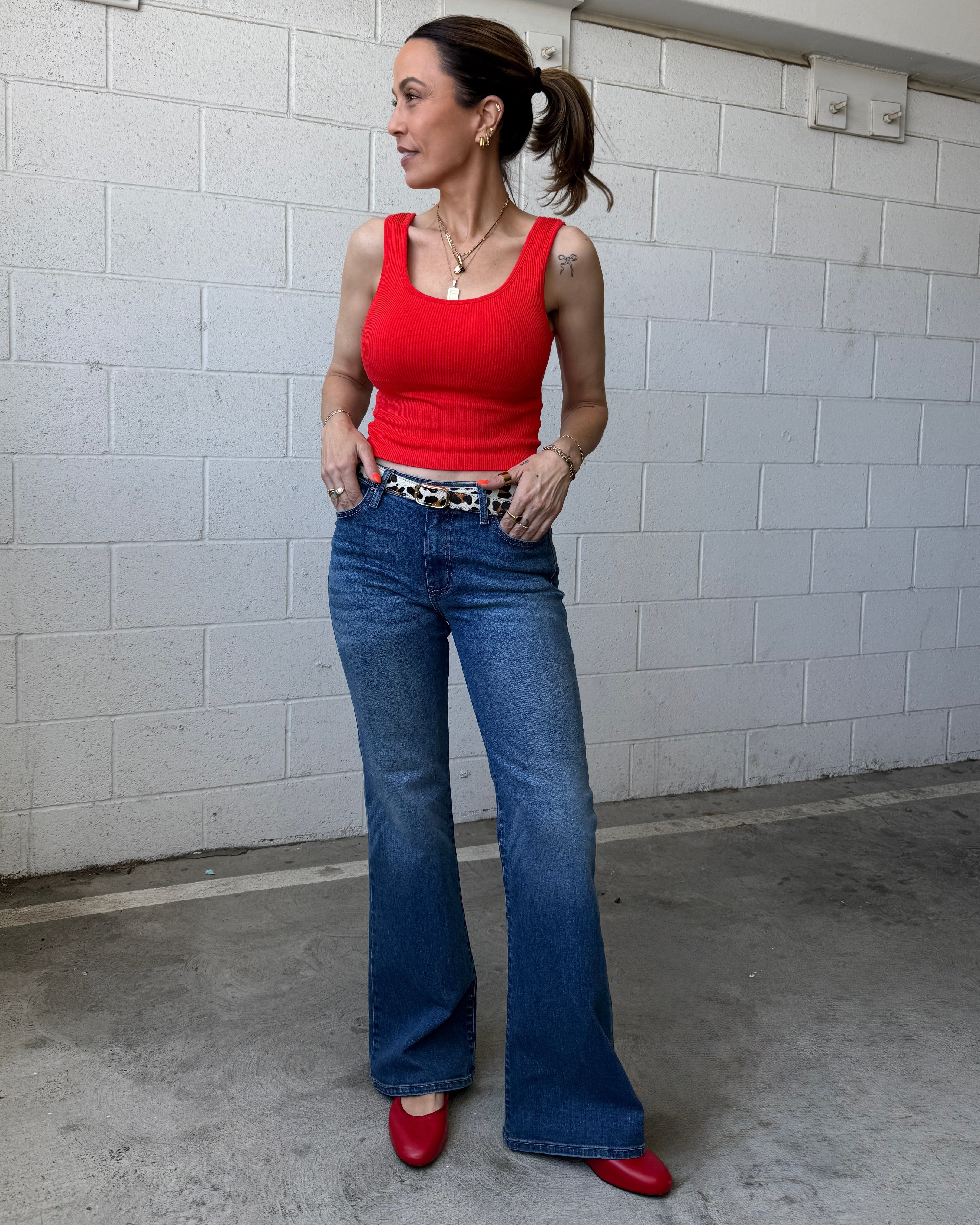 Woman wearing a red tank top and blue jeans standing against a white tiled wall.
