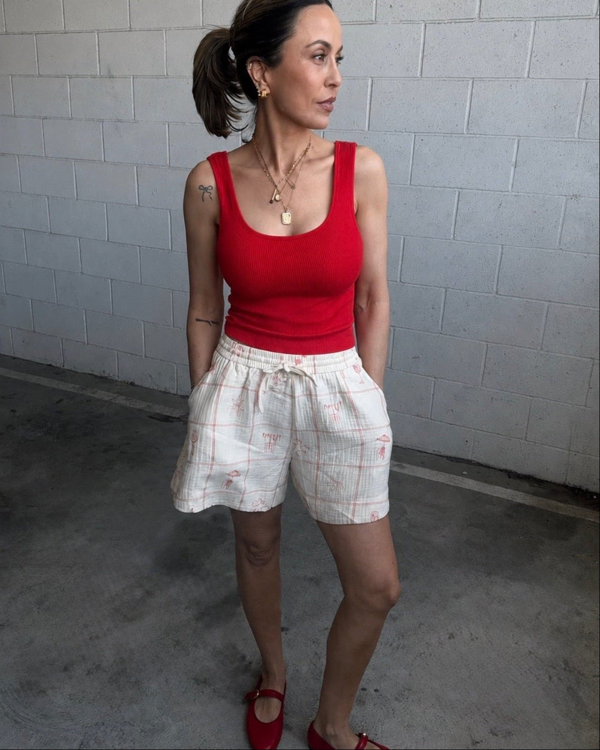 Woman wearing a red tank top and white plaid shorts standing against a gray brick wall.
