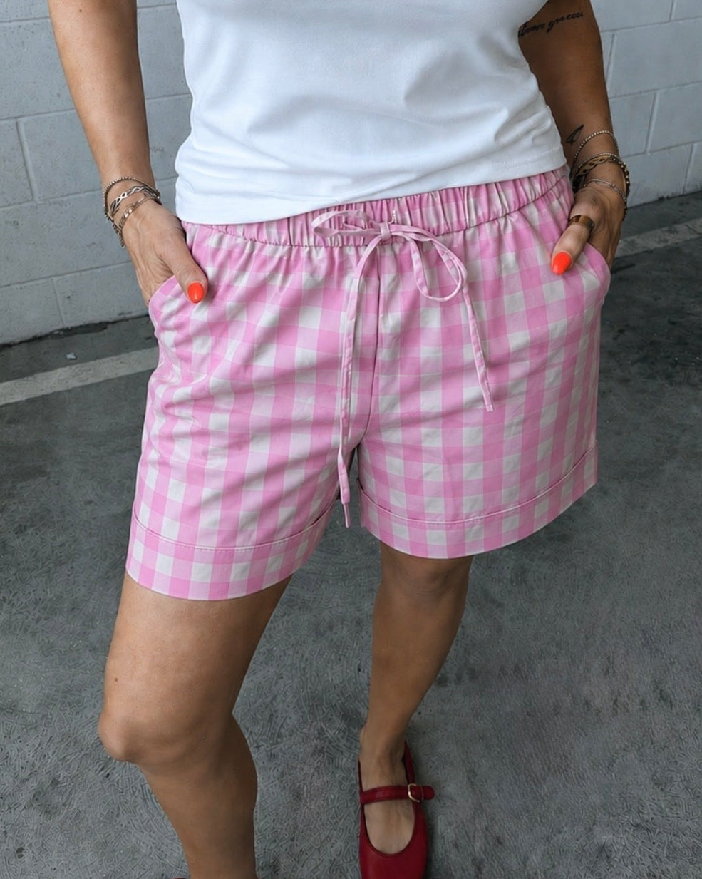 Person wearing a white top and pink checkered shorts with red shoes, standing against a tiled wall.