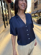 Woman standing outside wearing silky short sleeve, navy blue collared blouse with shite button. 