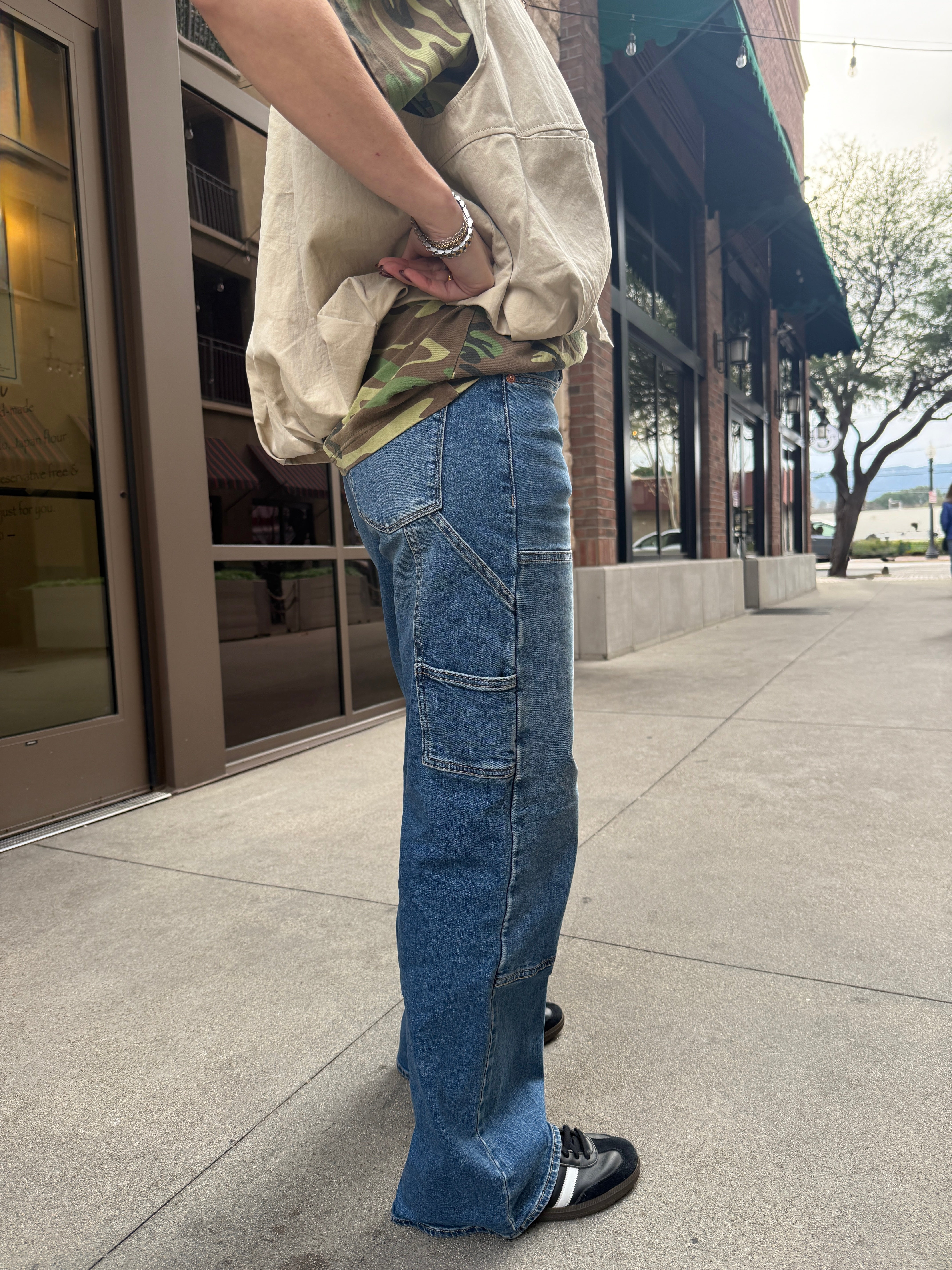 Woman wearing carpenter jeans in urban setting with tan vest and camo t-shirt