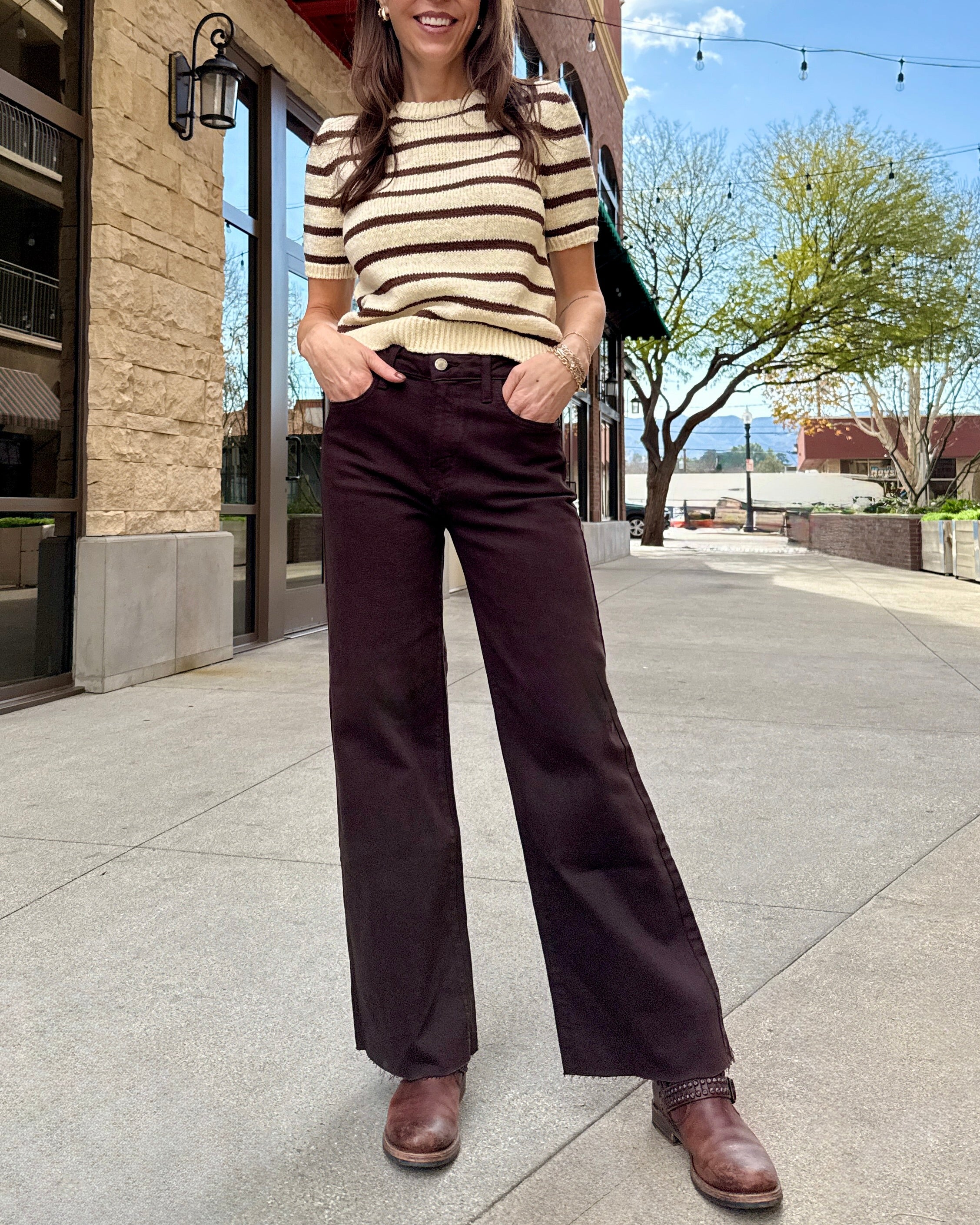 Woman standing outside wearing brown raw hem jeans with stripe cream and brown sweater. 