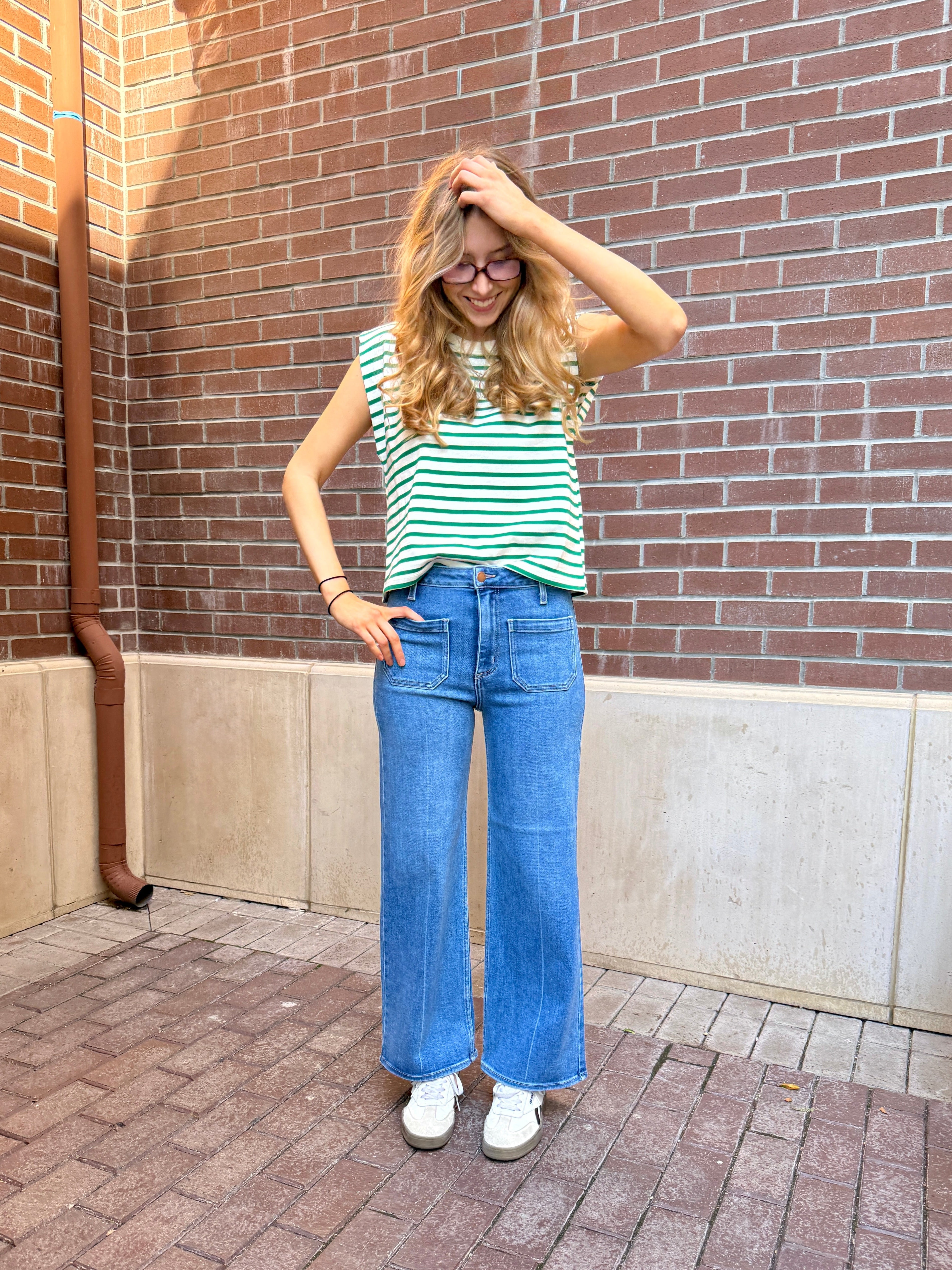 Girl in front of red brick wall, with hand on her head, wearing jeans with front pockets and black and white striped cap sleeve t-shirt. 