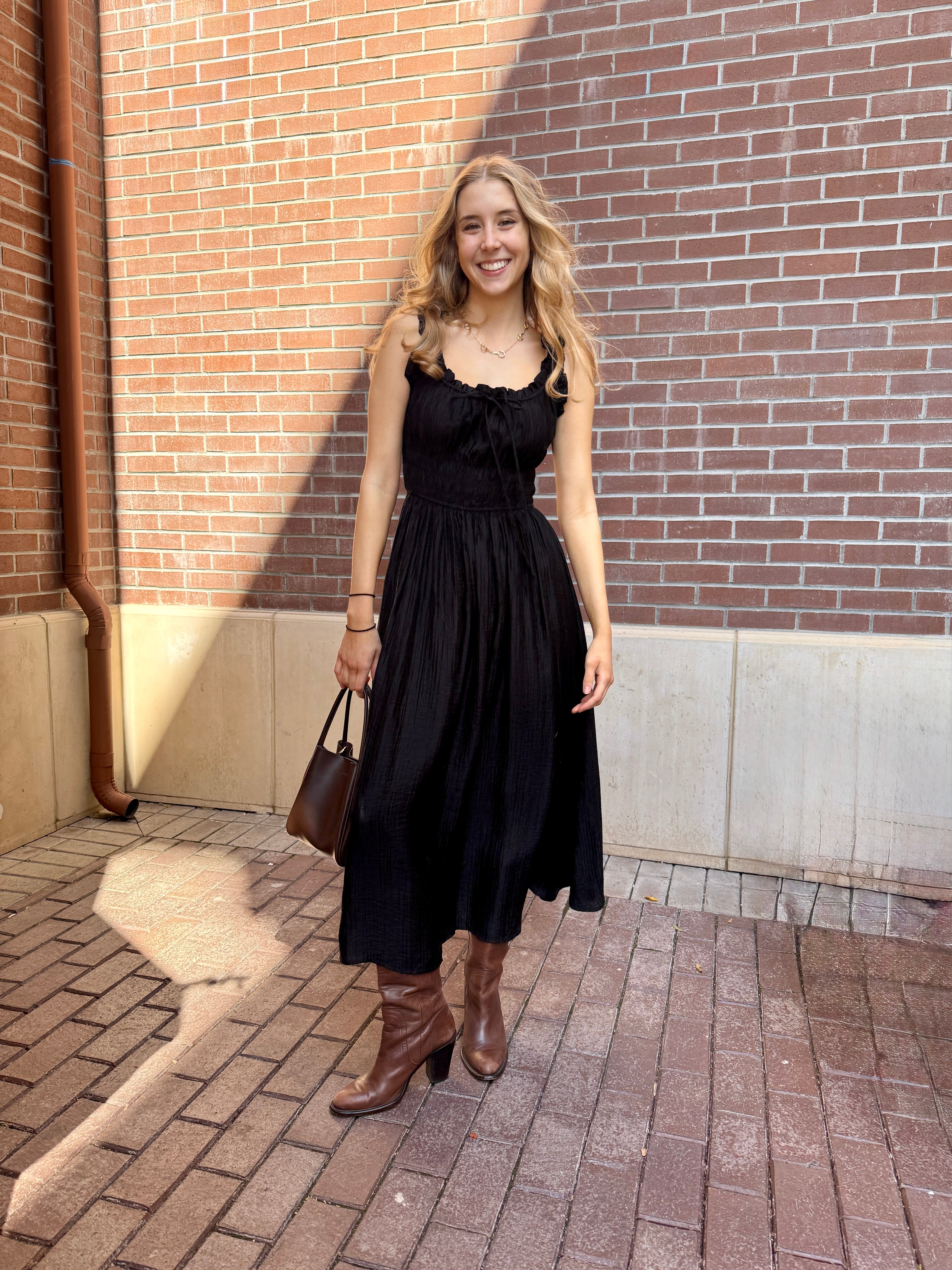 Model outside against a brick building wearing long black dress with thick straps and brown boots 