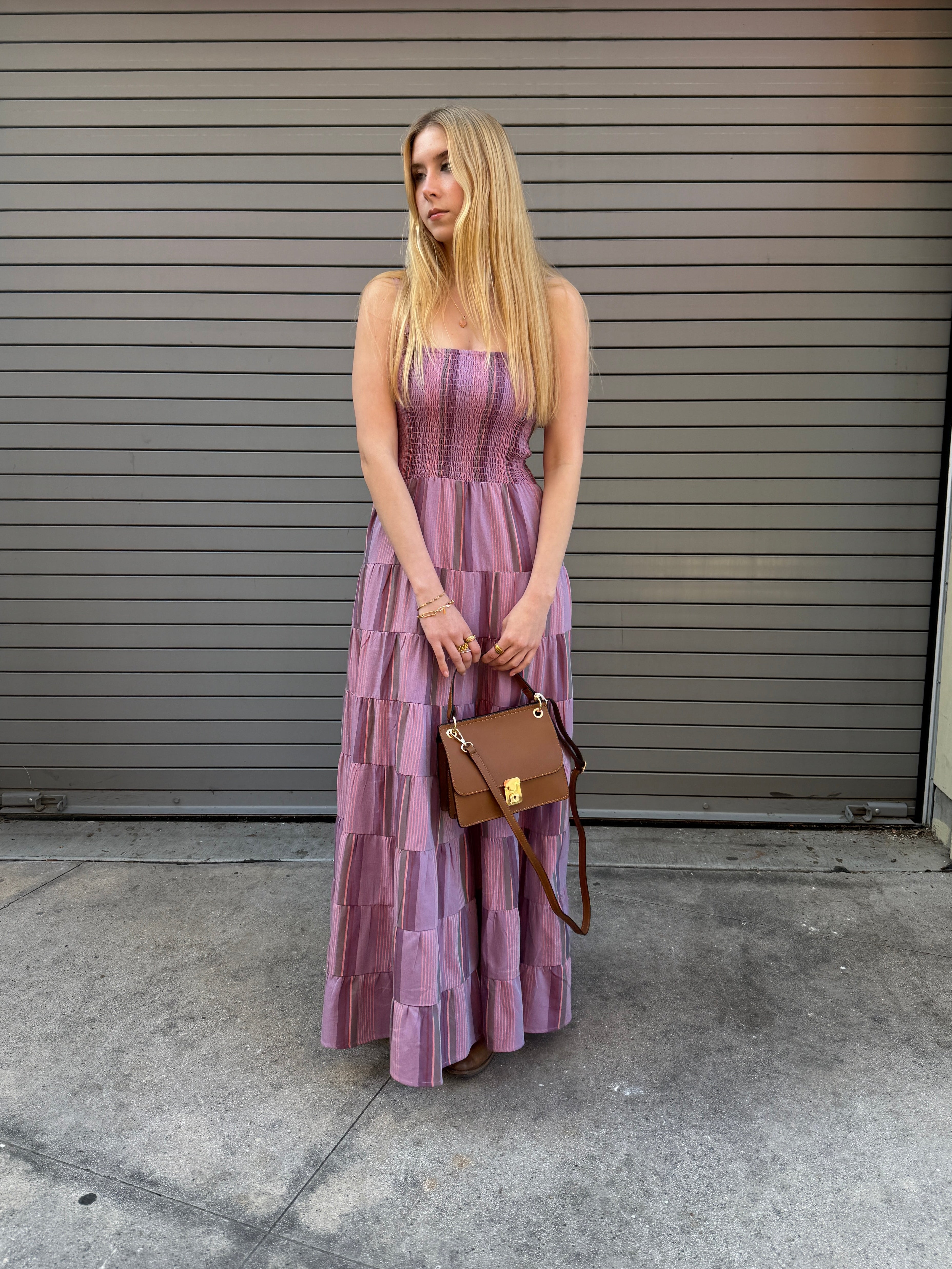 Woman in a purple dress holding a brown handbag against a gray wall.