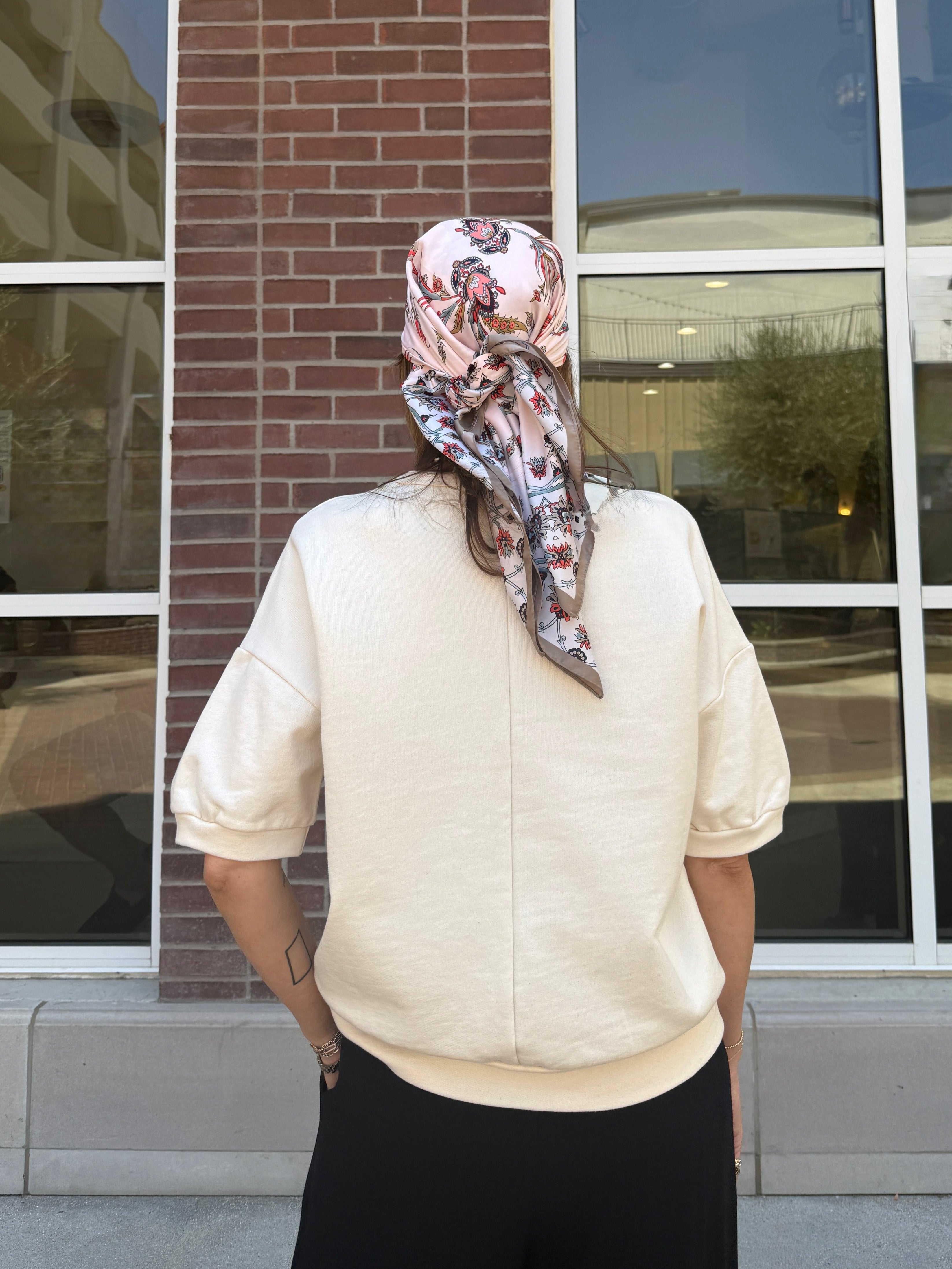 Person wearing a patterned headscarf standing in front of a building with large windows With cream short sleeve sweatshirt