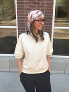 Woman wearing a headscarf and glasses standing in front of a brick wall with cream short sleeve sweatshirt 