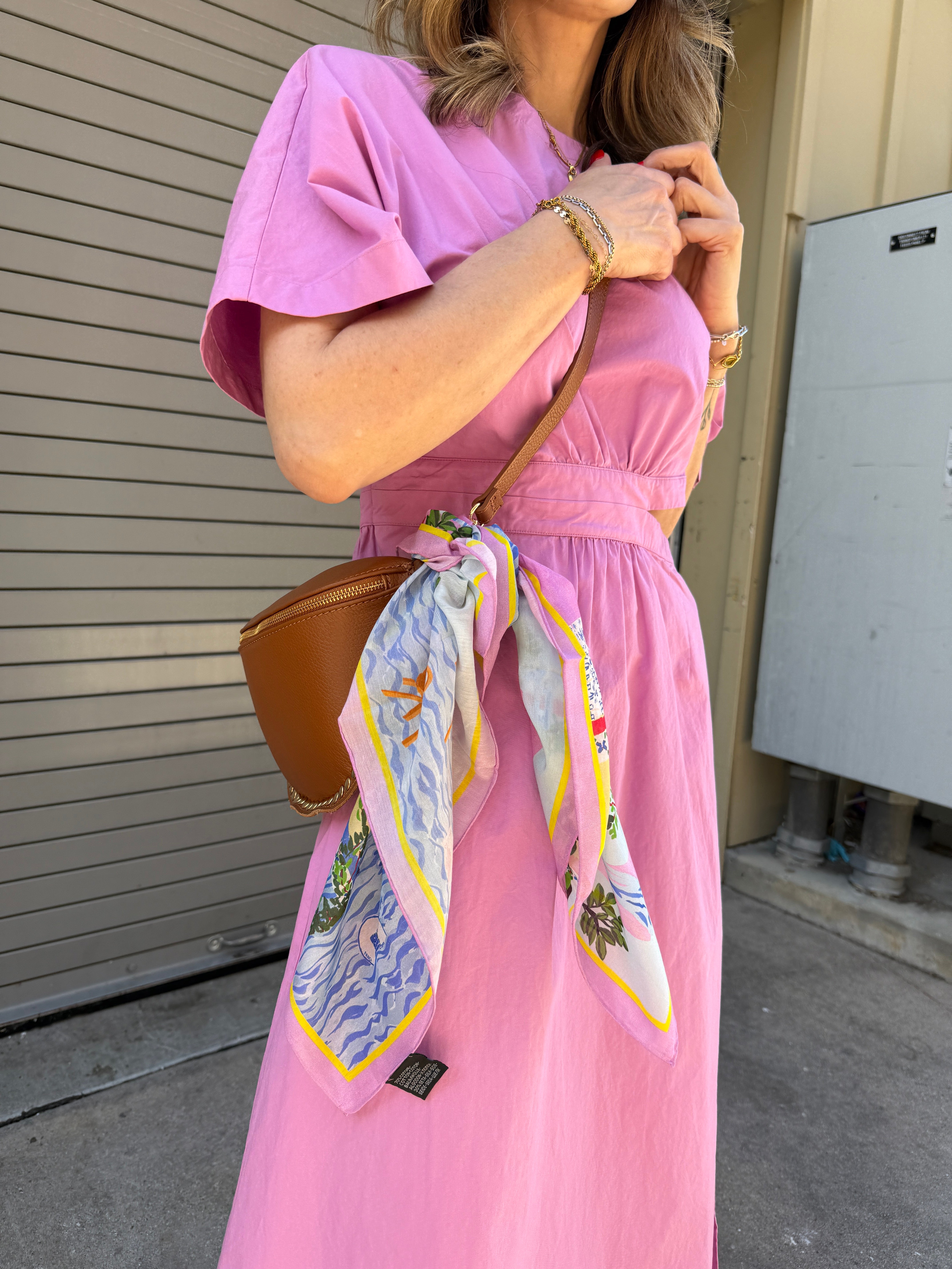 Person wearing a pink dress with a colorful scarf and holding a brown bag on a deck.