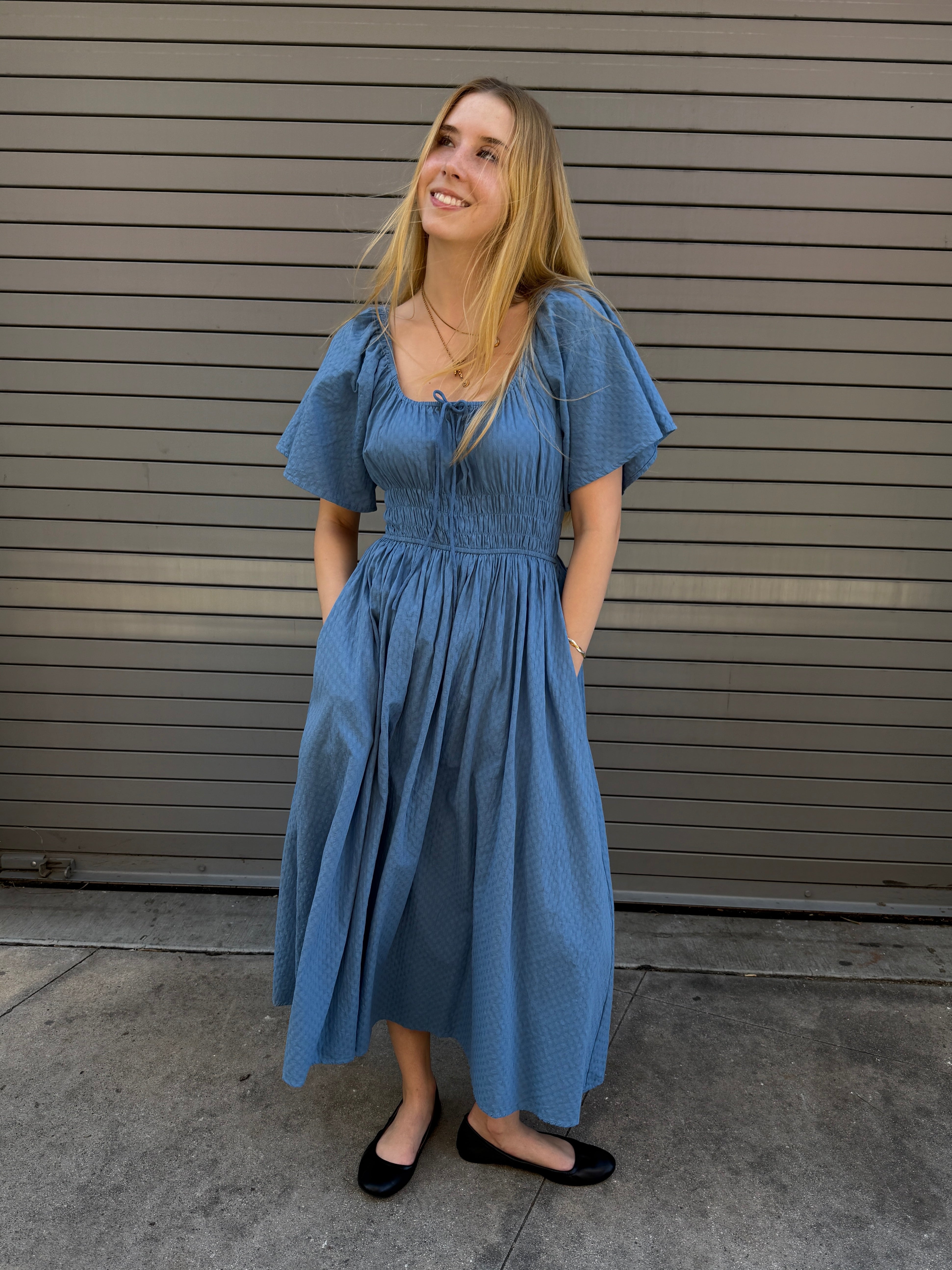 Woman wearing a blue dress standing against a gray wall.