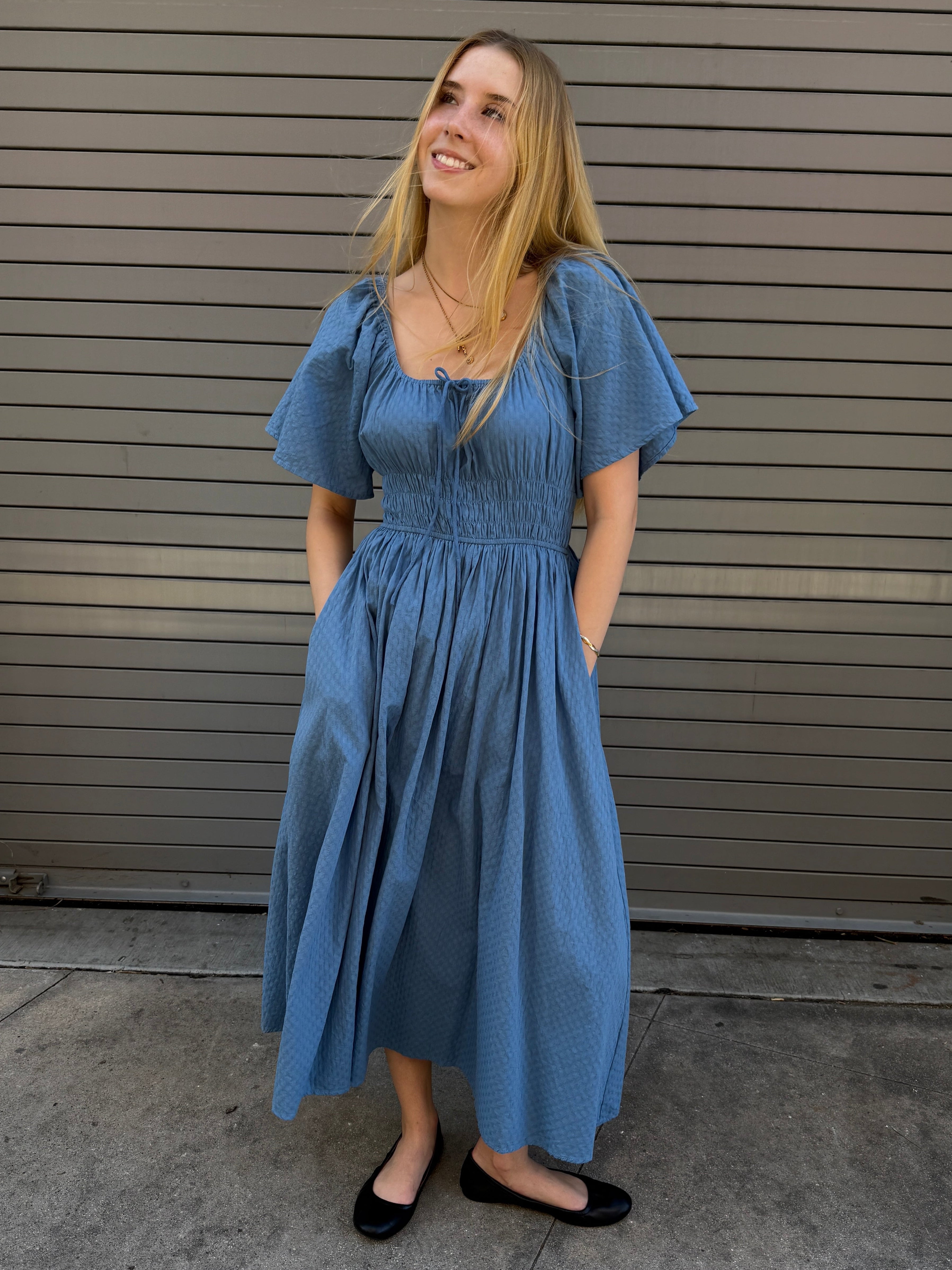 Woman wearing a blue dress standing against a gray wall.