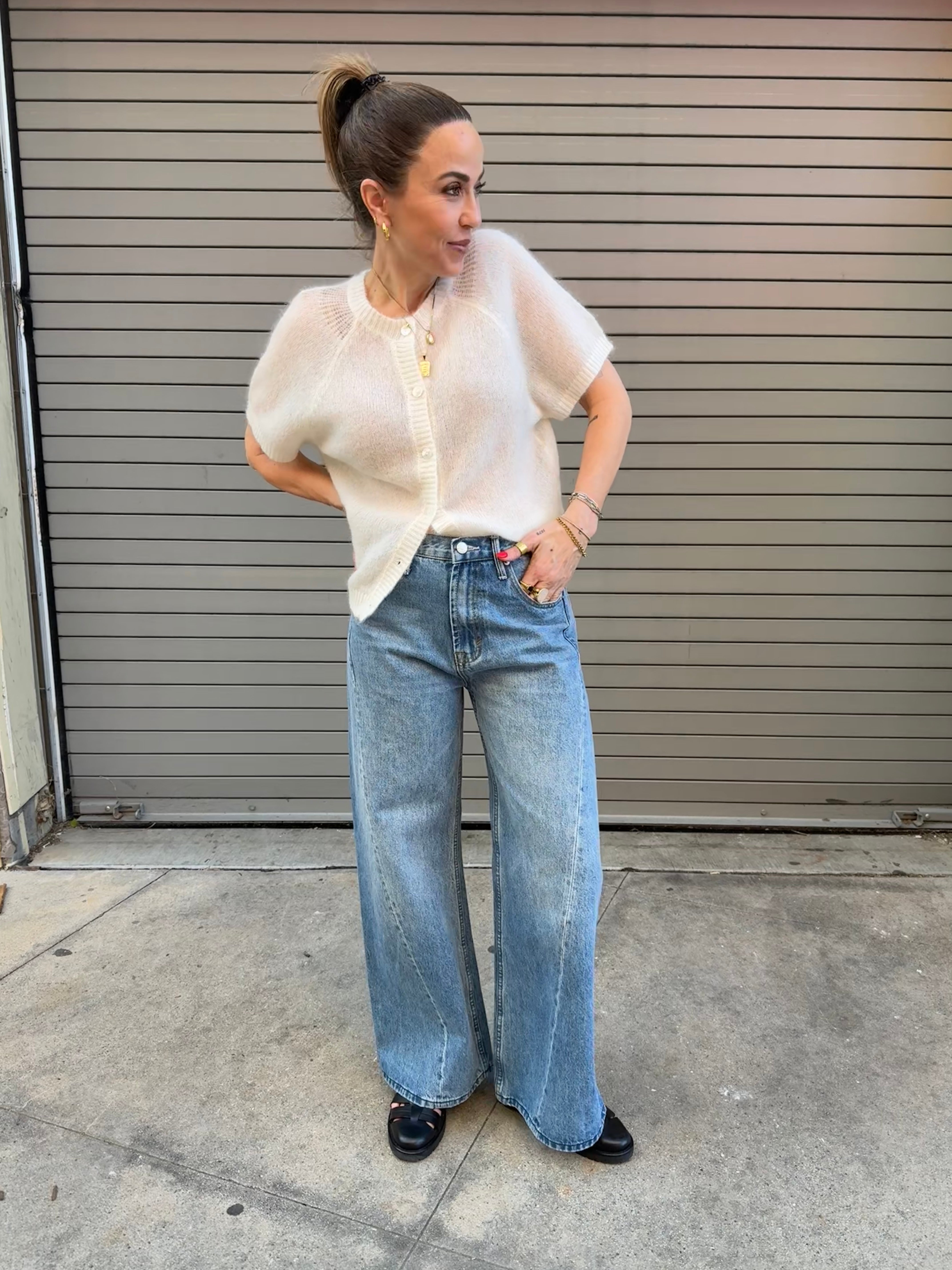 Woman wearing a sheer white sweater and blue jeans standing in front of a closed garage door.
