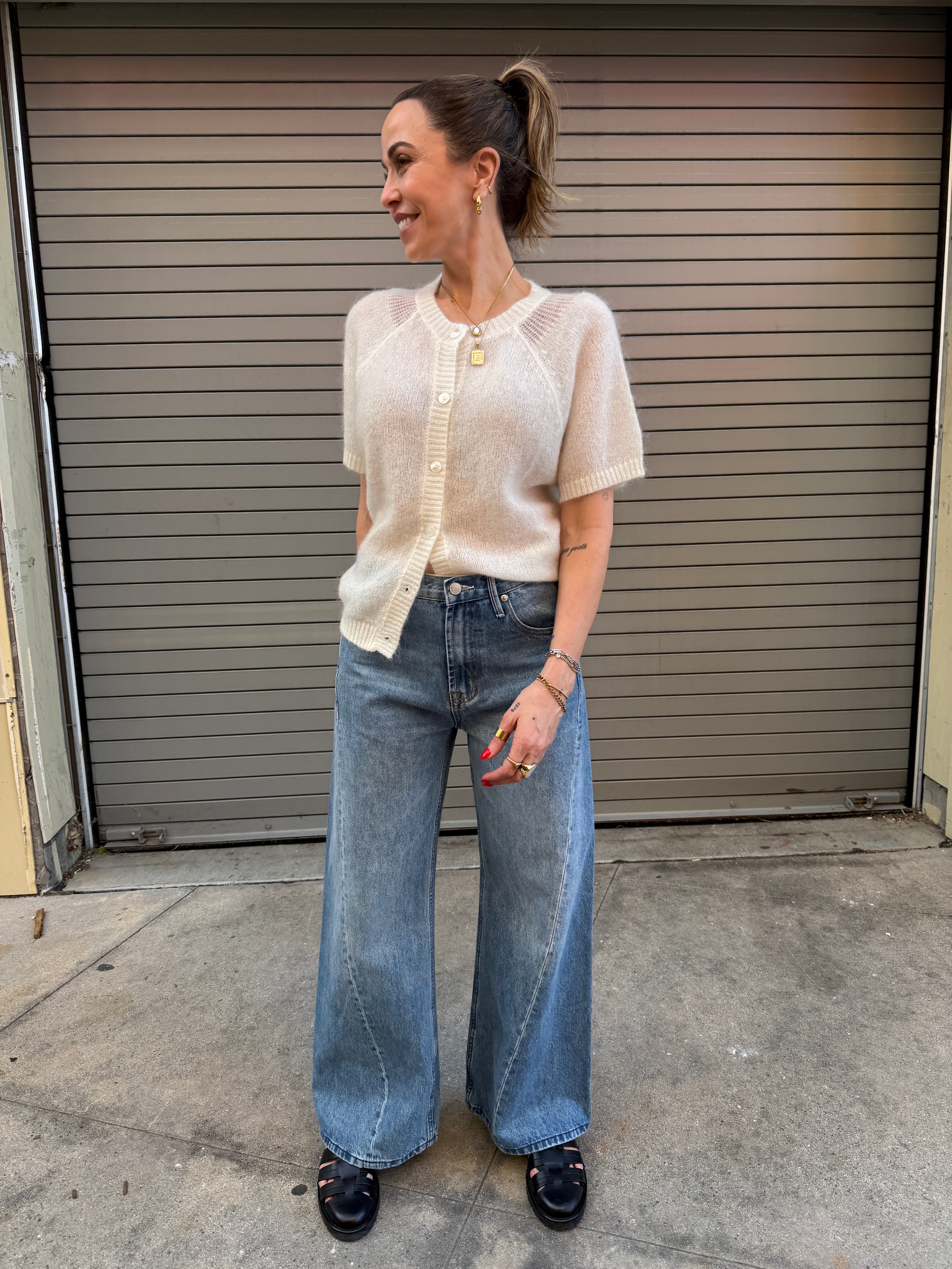 Woman wearing a beige vest and blue jeans standing on a sidewalk.