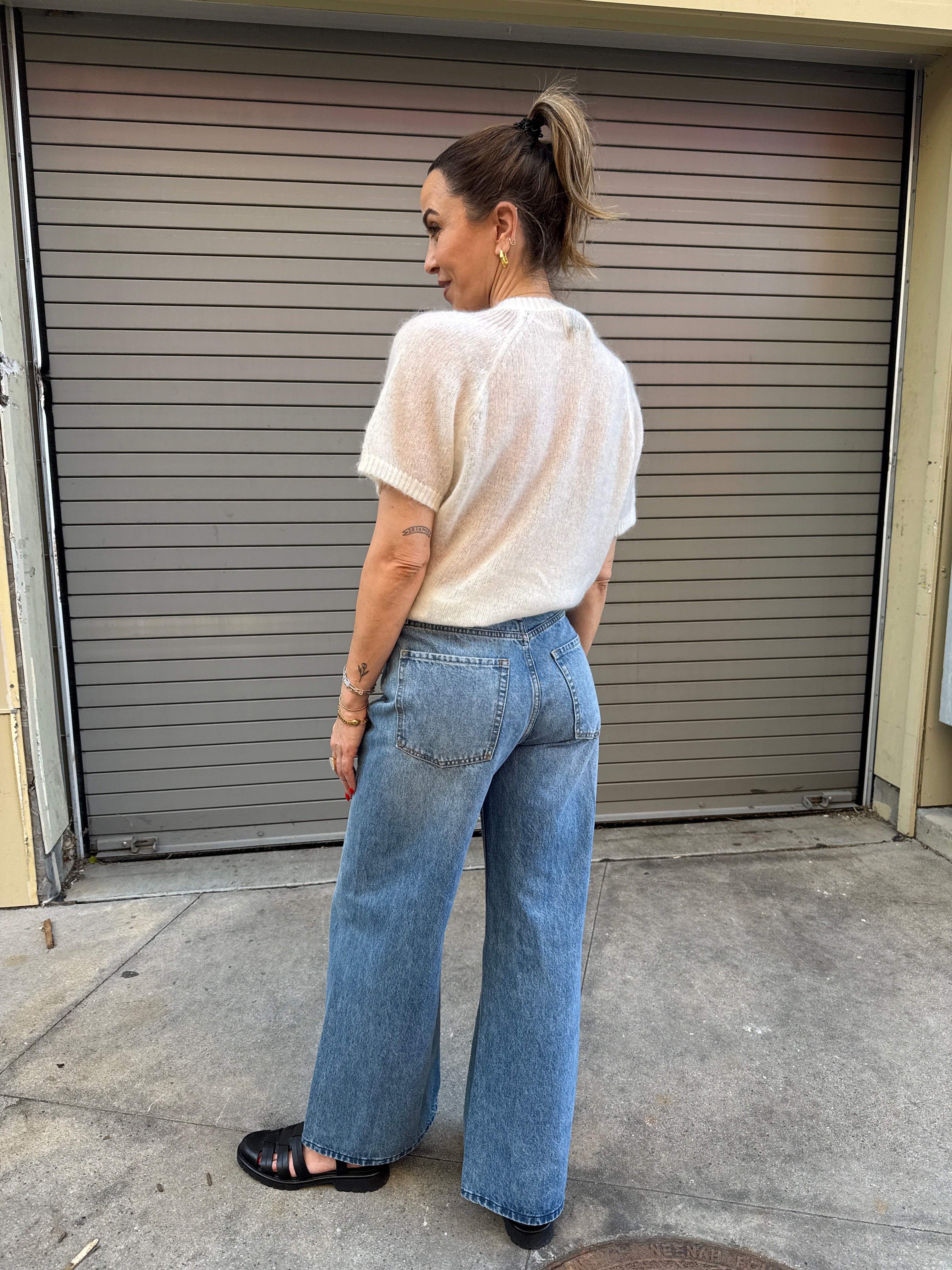 Person wearing a light-colored sweater and blue jeans standing in front of a garage door.