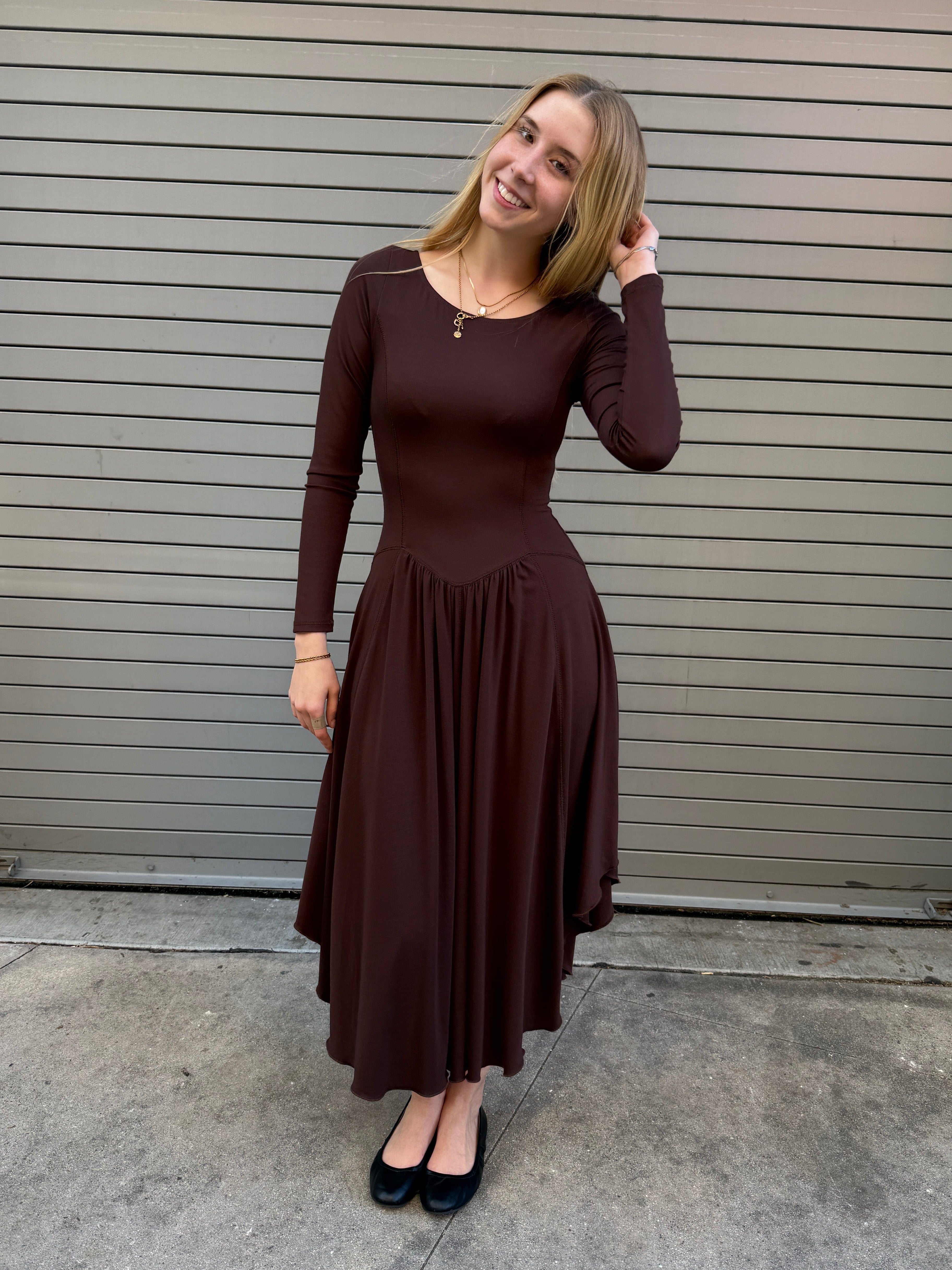Woman in a brown dress standing on a sidewalk with a building in the background