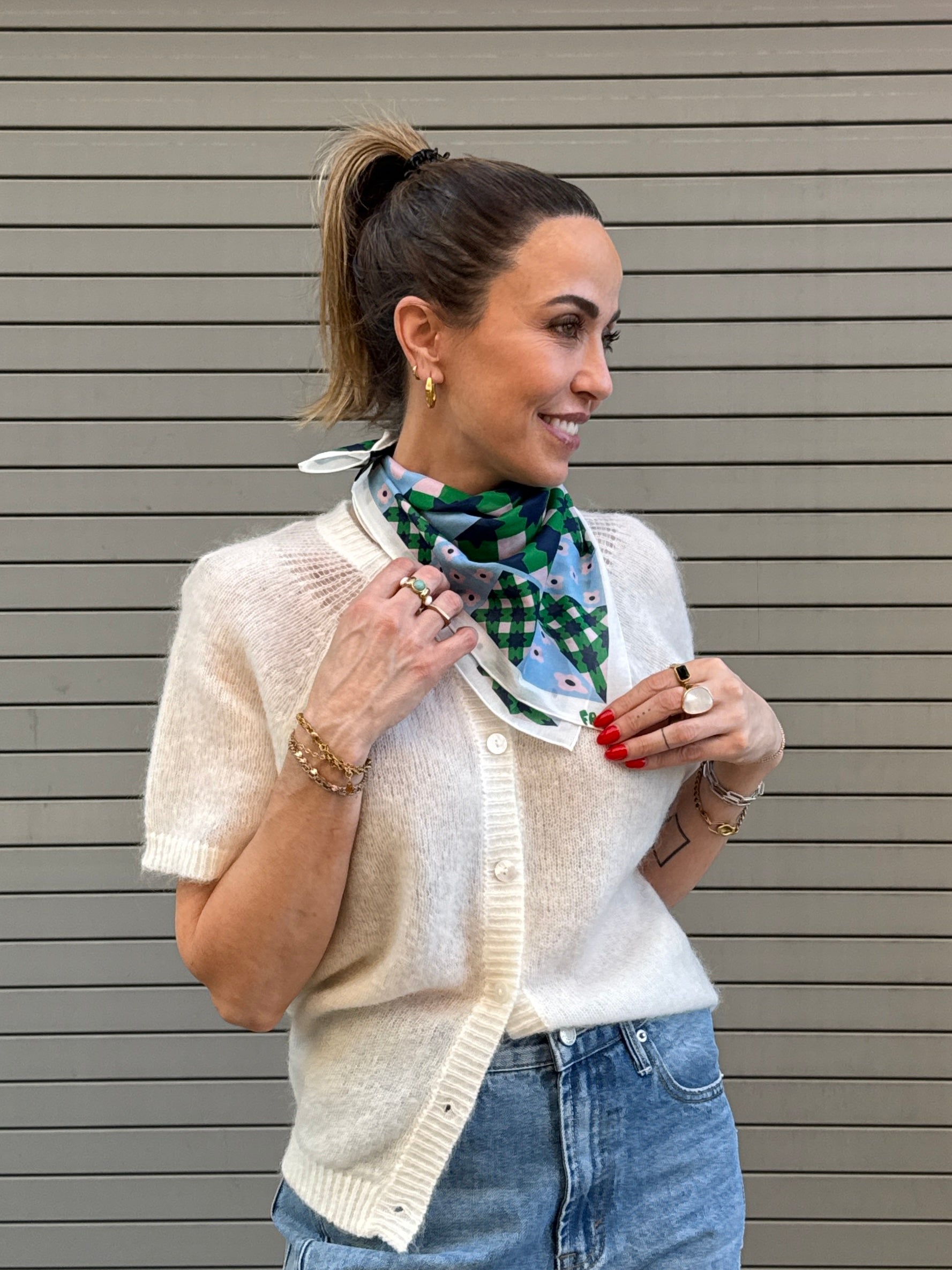 Woman wearing a white cardigan, blue jeans, and a patterned scarf against a gray background