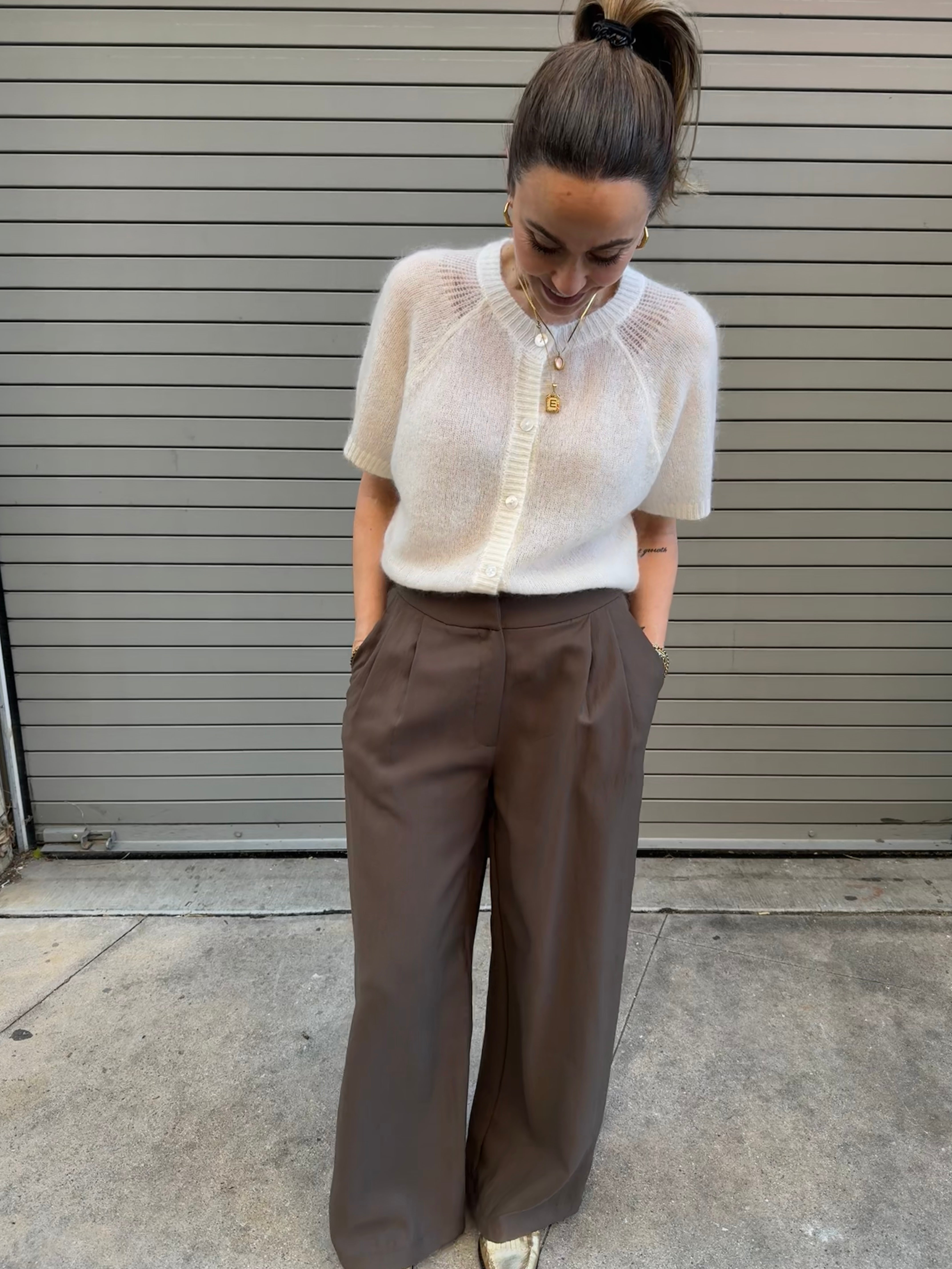 Woman wearing a white knit top and brown pants standing in front of a gray wall.