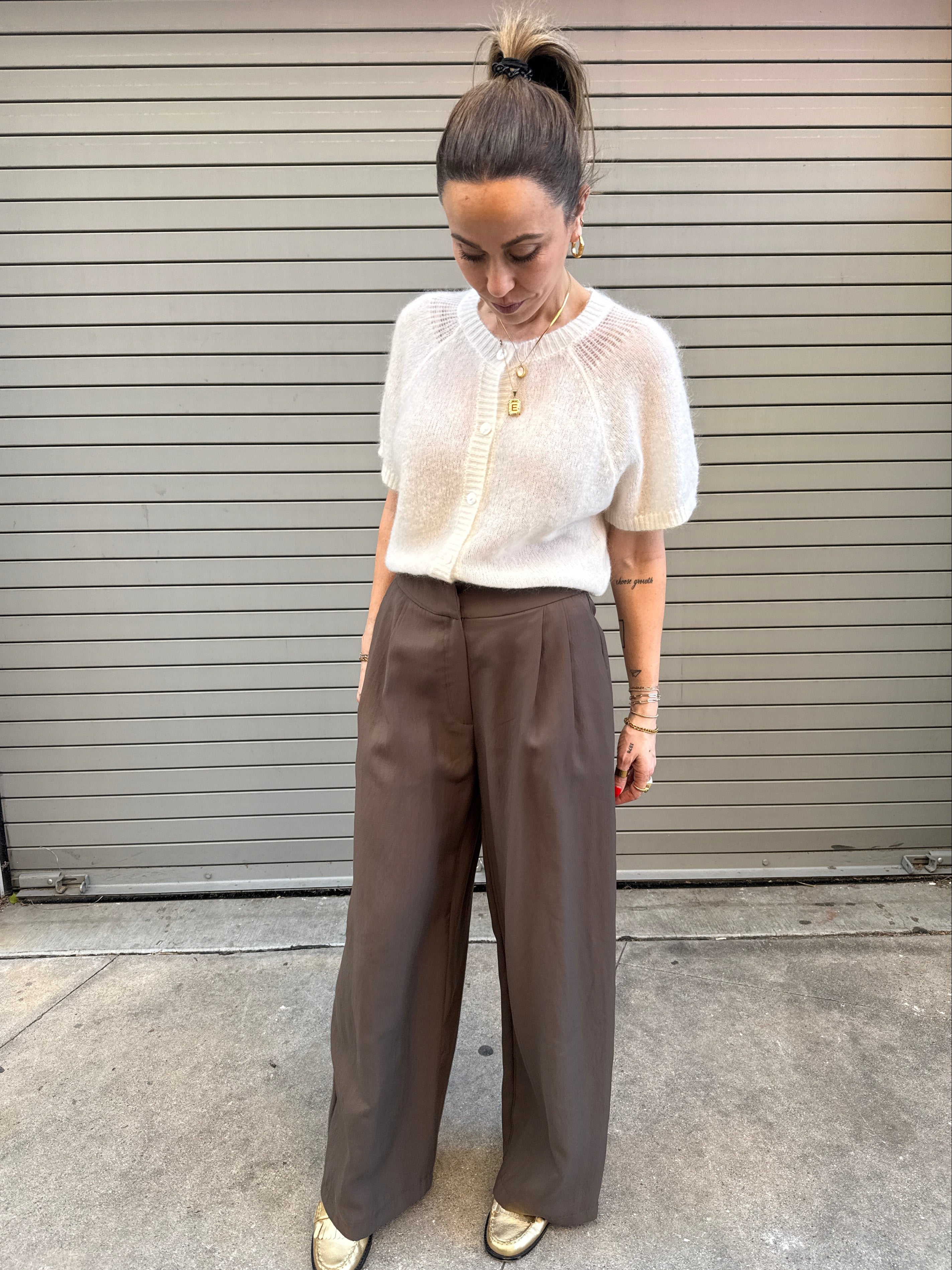 Person wearing a white sweater and brown pants standing against a gray wall.