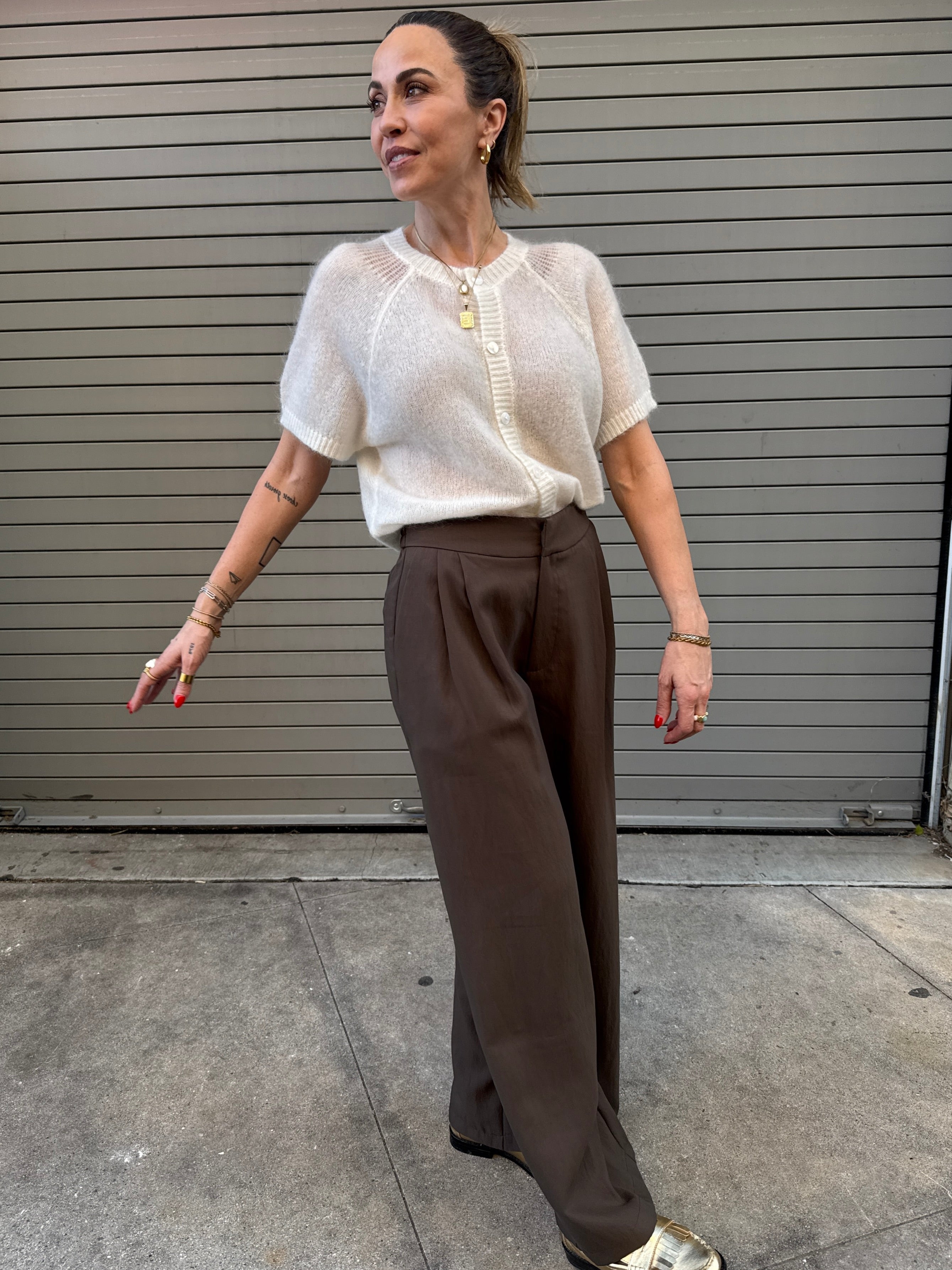Woman wearing a white top and brown pants standing in front of a gray metal shutter.