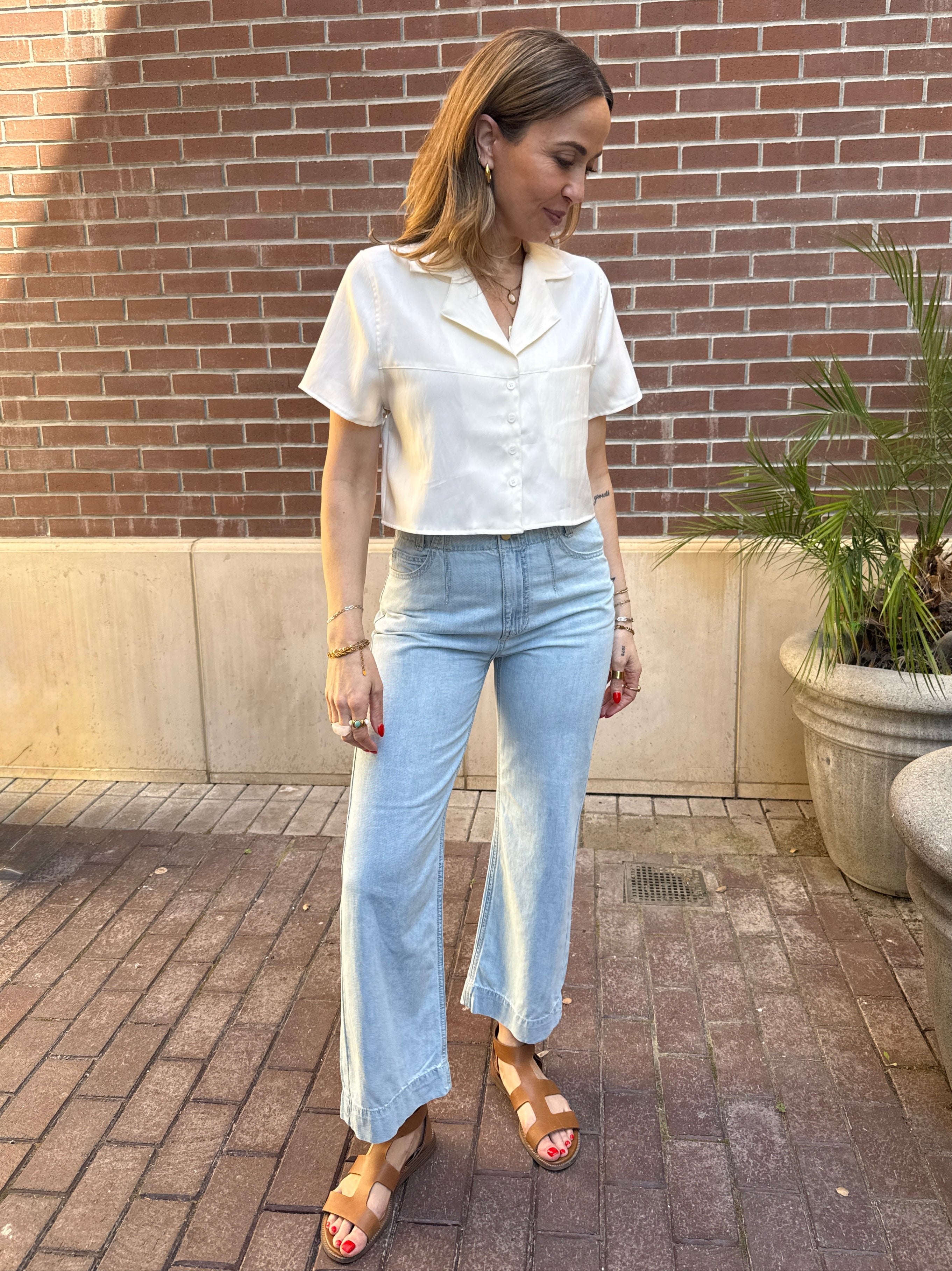 Woman in a white blouse and light blue jeans standing against a brick wall.