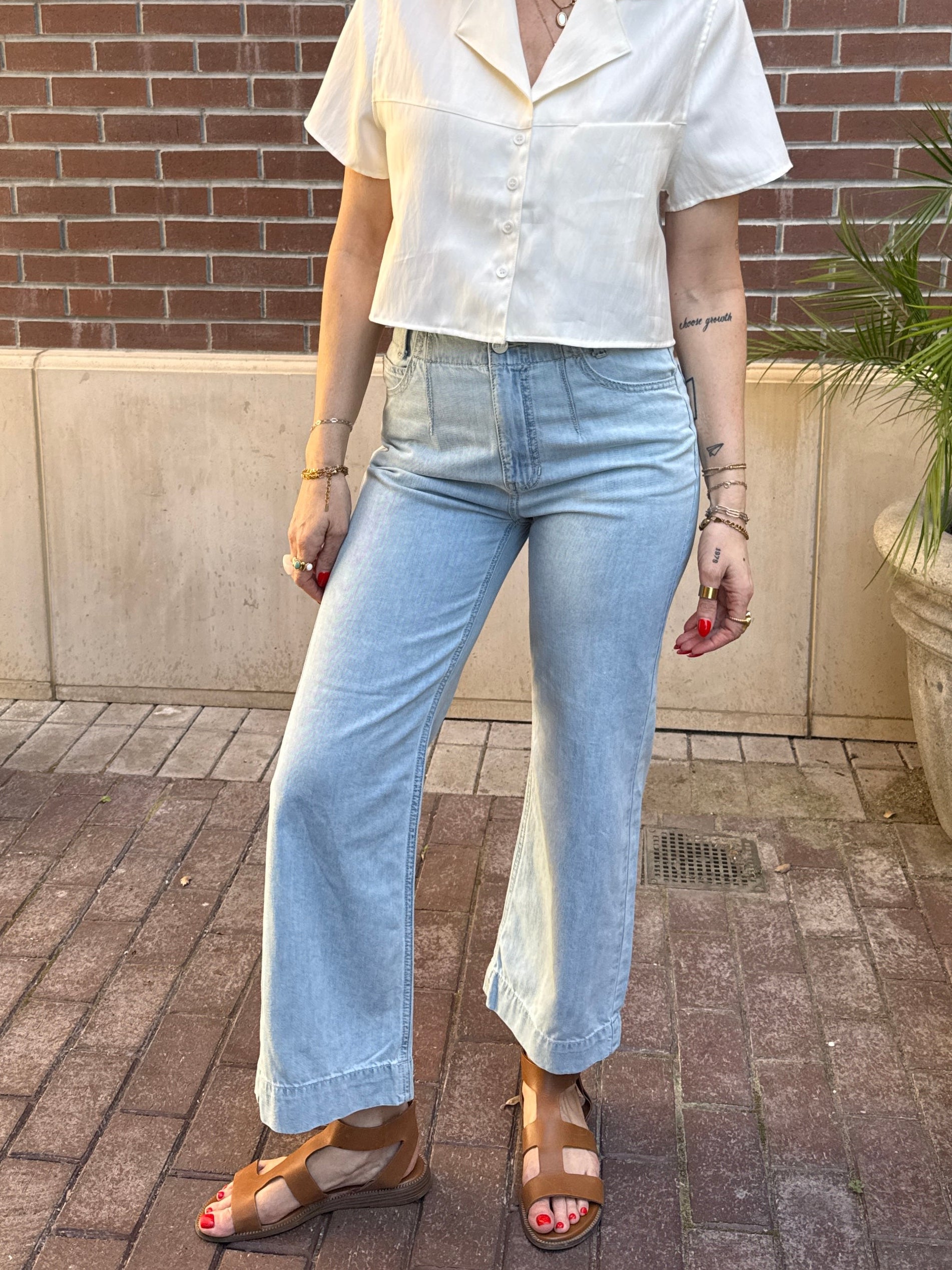 Person wearing a white shirt and light blue jeans standing on a sidewalk with a brick wall and plant in the background.