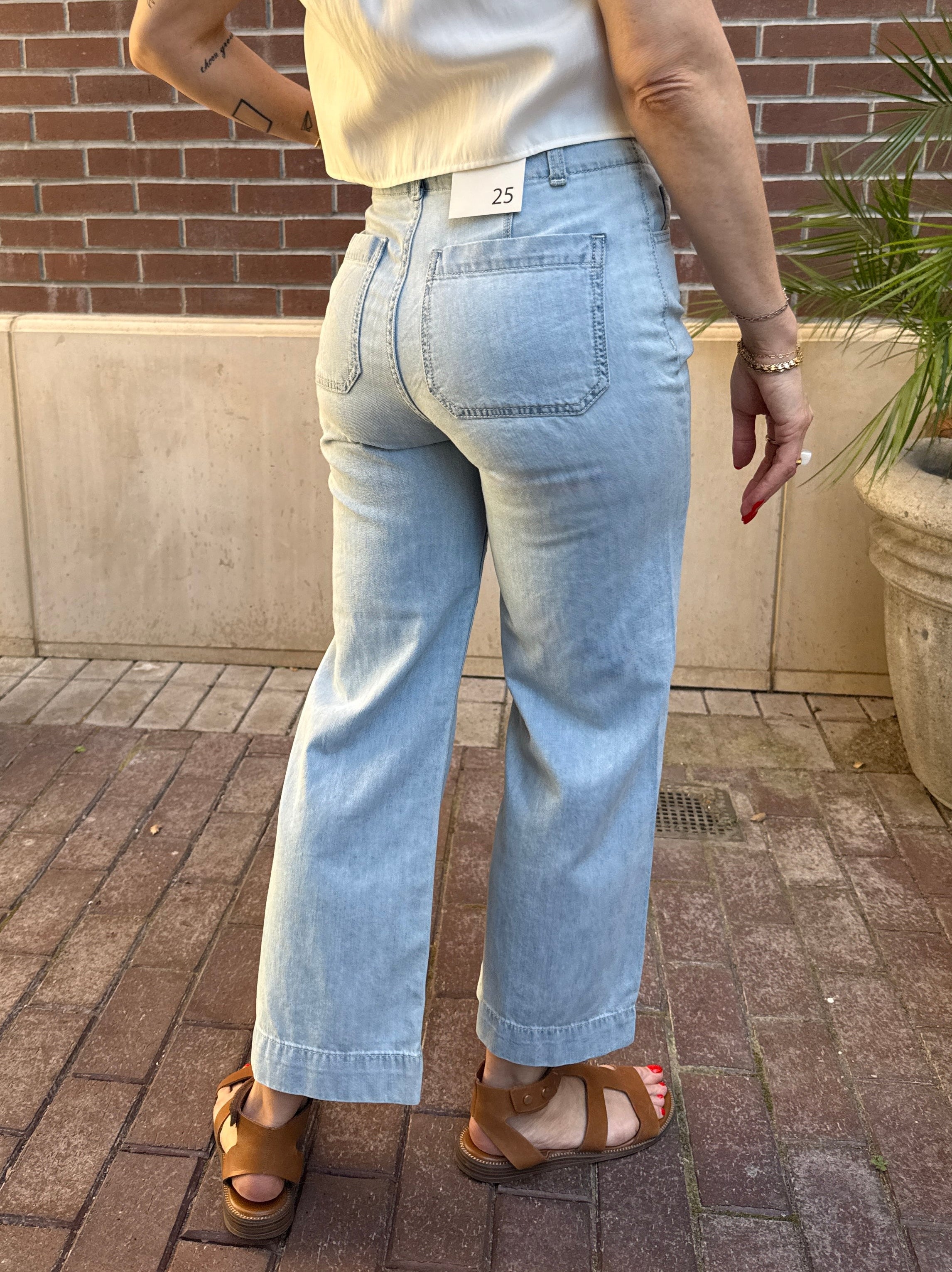 Person wearing light blue jeans standing on a patio with a brick wall and potted plant in the background.