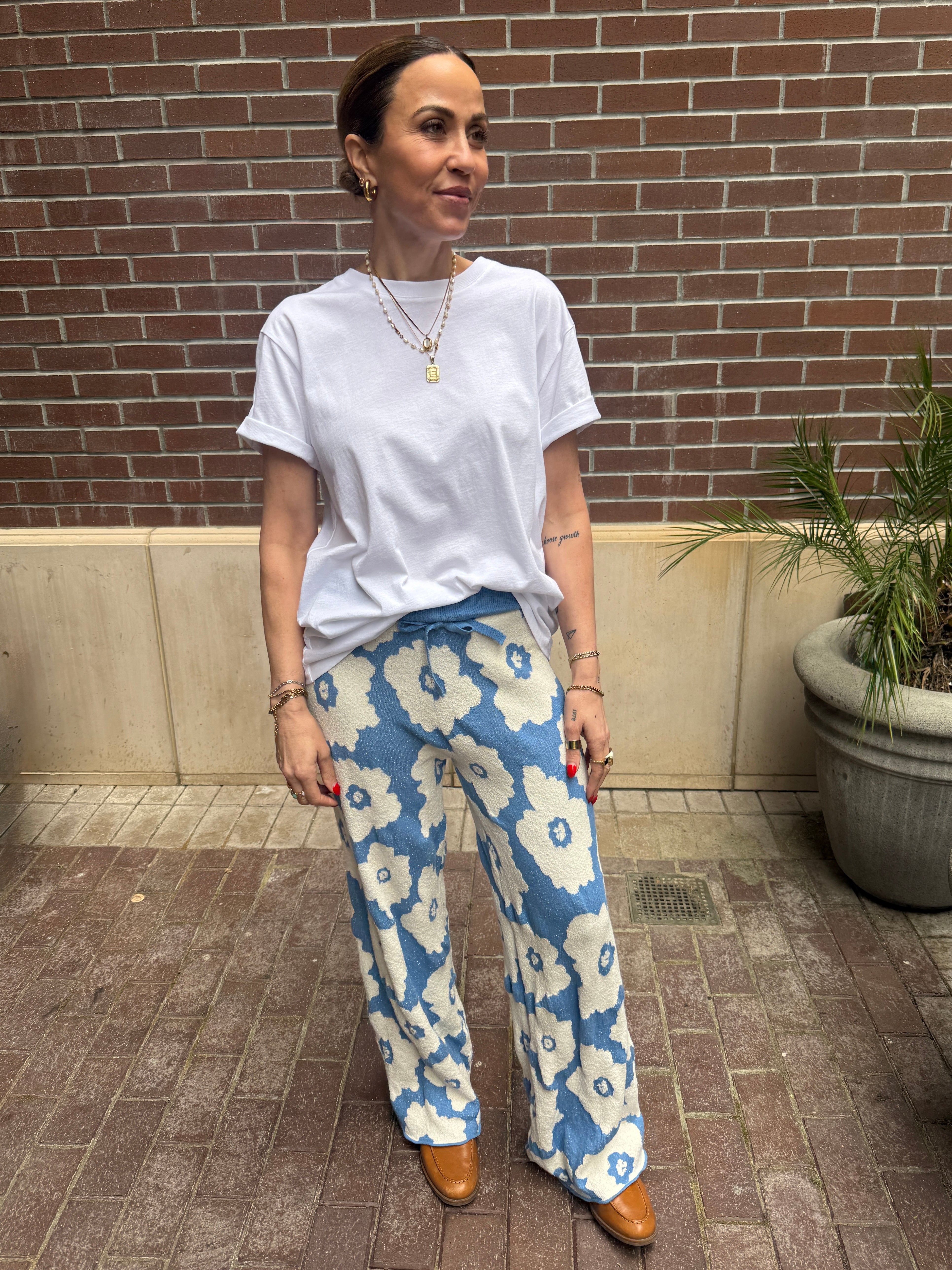 Woman wearing a white t-shirt and blue floral pants standing against a brick wall.