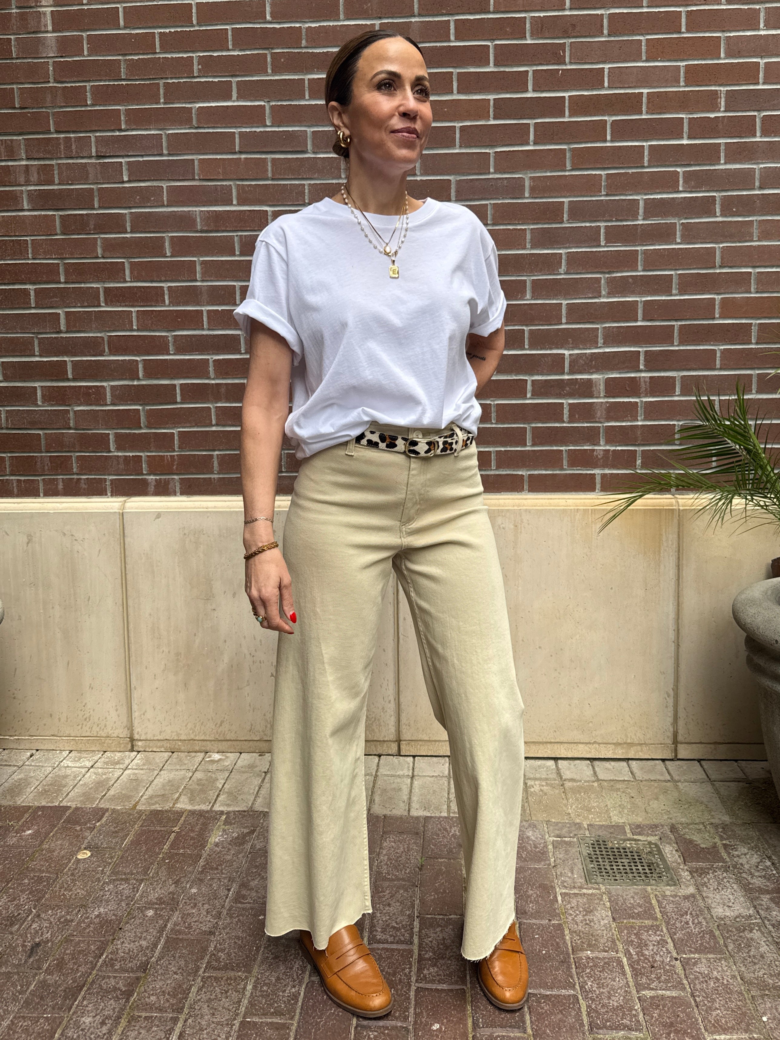Woman wearing a white t-shirt and beige pants standing against a brick wall.