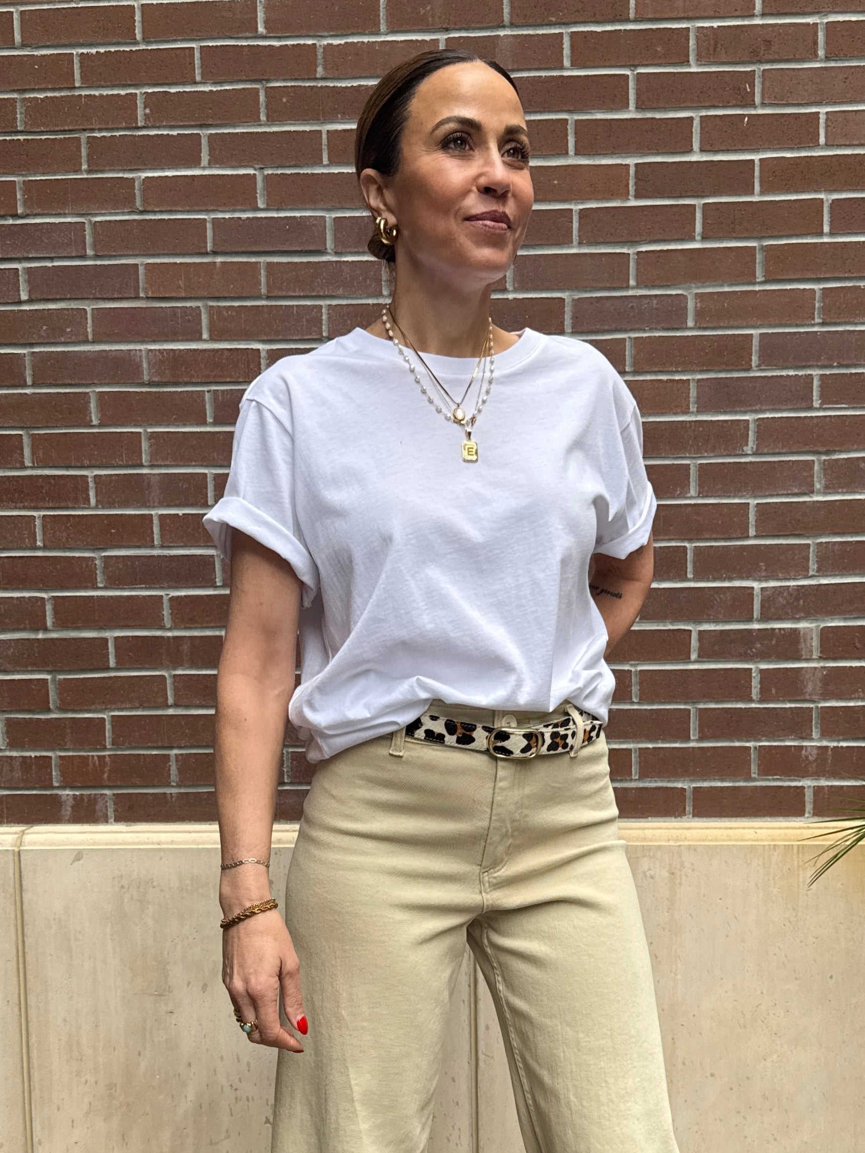 Woman wearing a white t-shirt and beige pants standing against a brick wall.