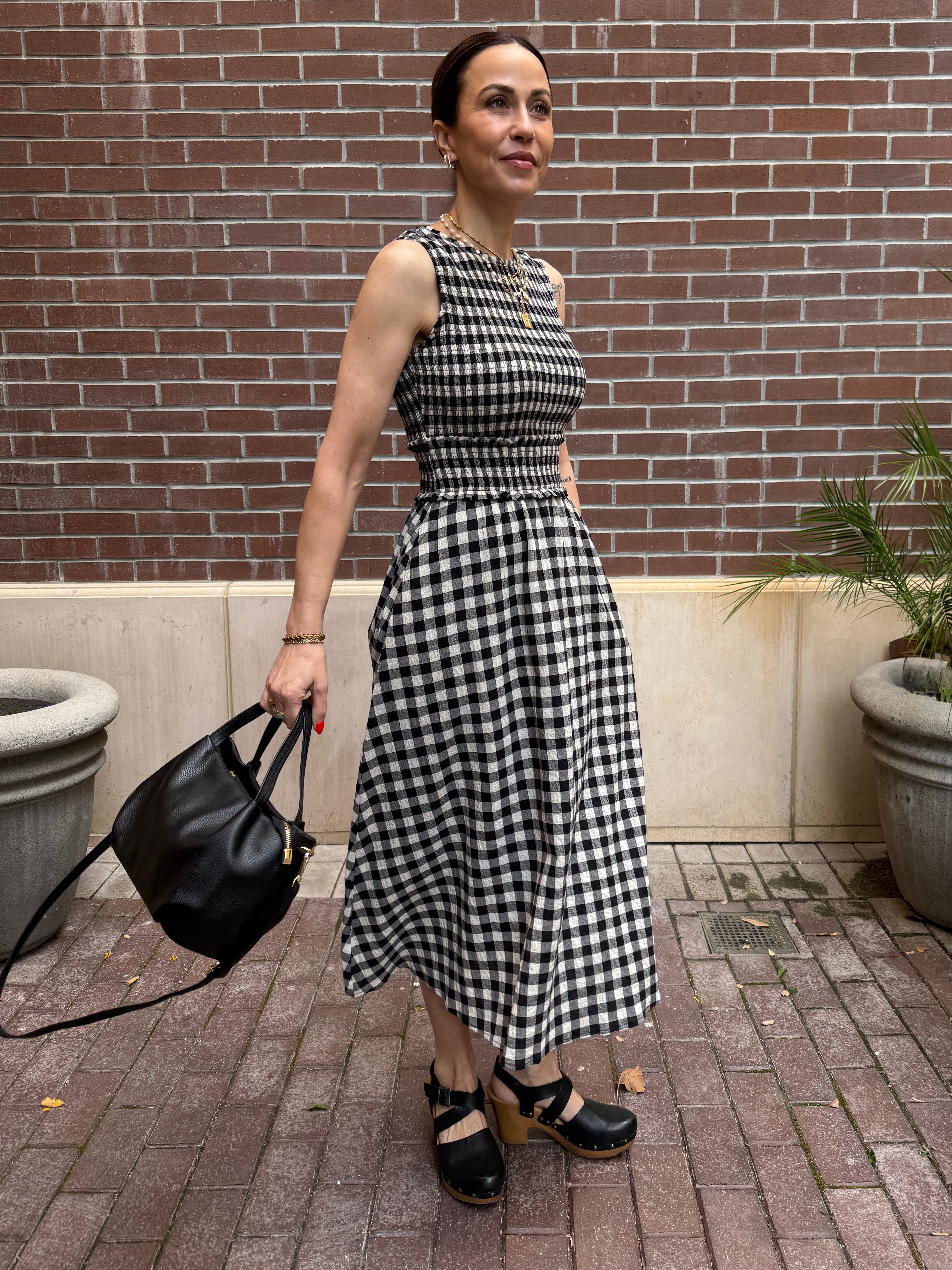 Woman in a black and white checkered dress holding a black bag against a brick wall.