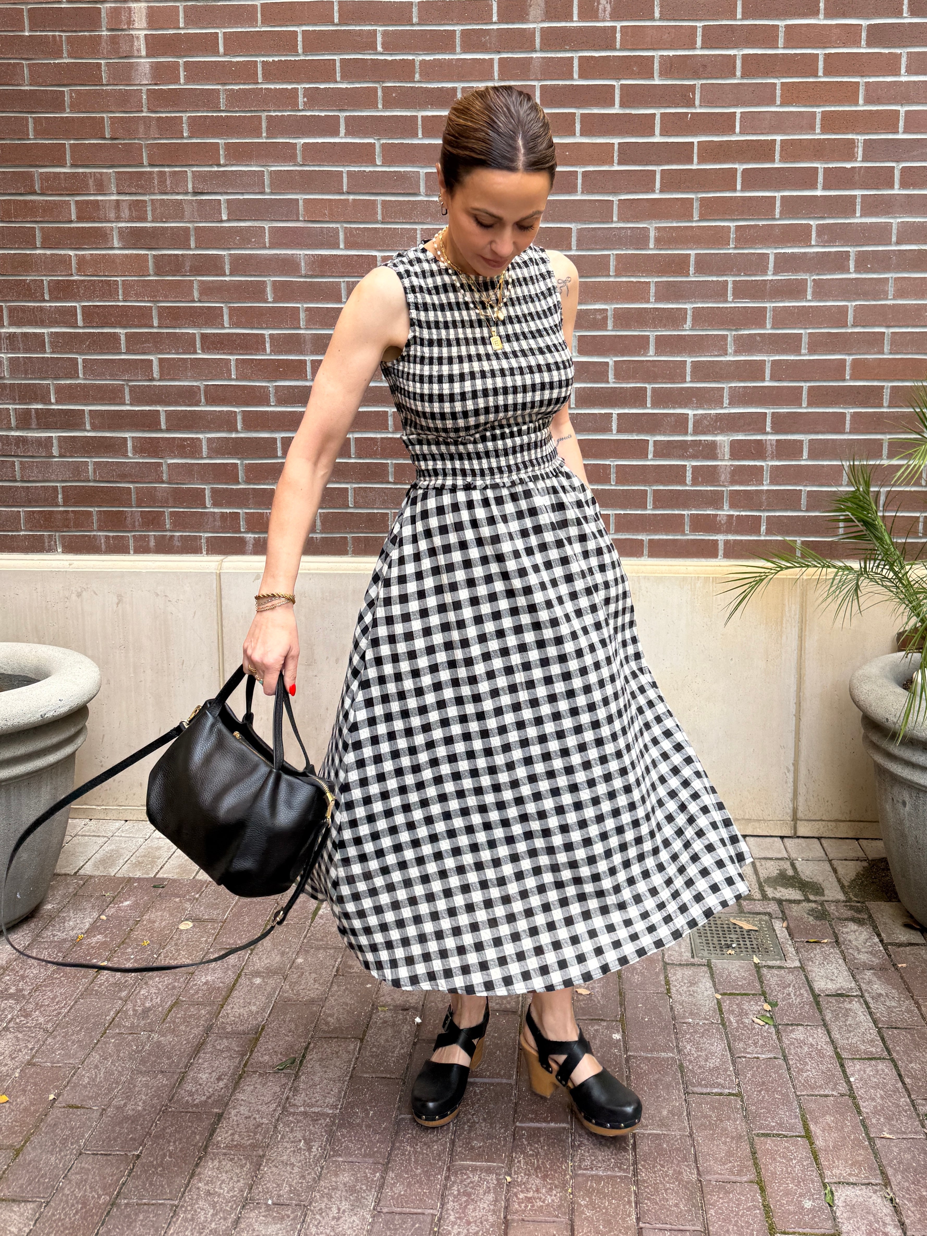 Woman in a black and white checkered dress holding a black handbag against a brick wall.