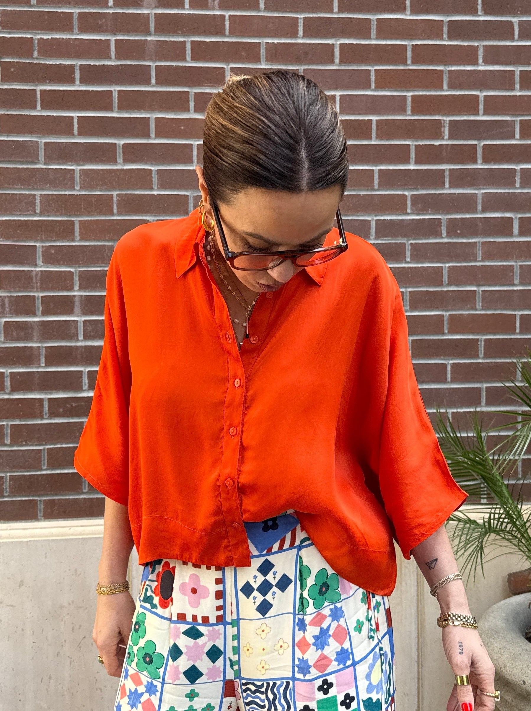 Person wearing an orange shirt and colorful pants standing against a brick wall.