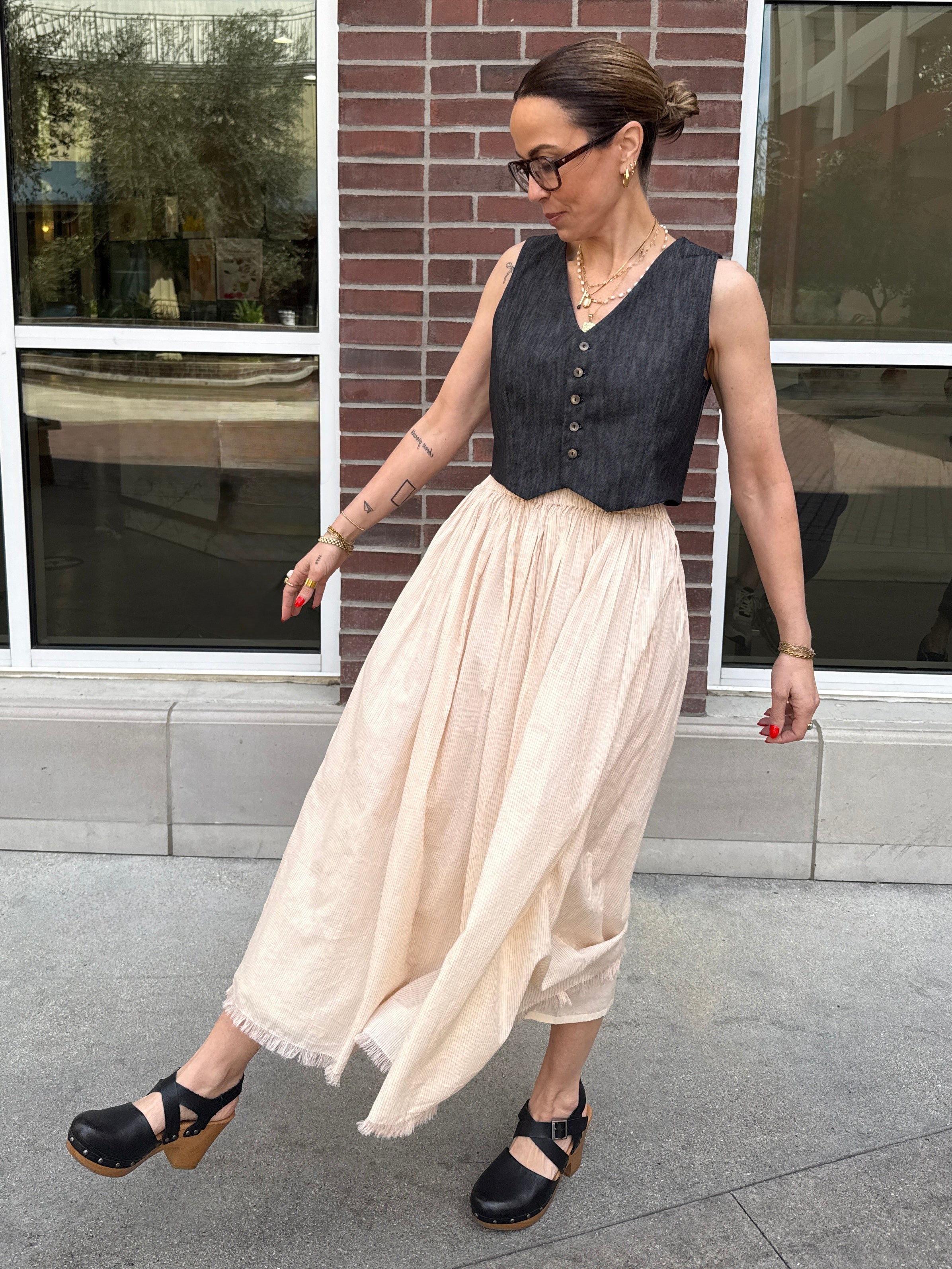 Woman wearing a sleeveless denim top and beige skirt standing outside a building.