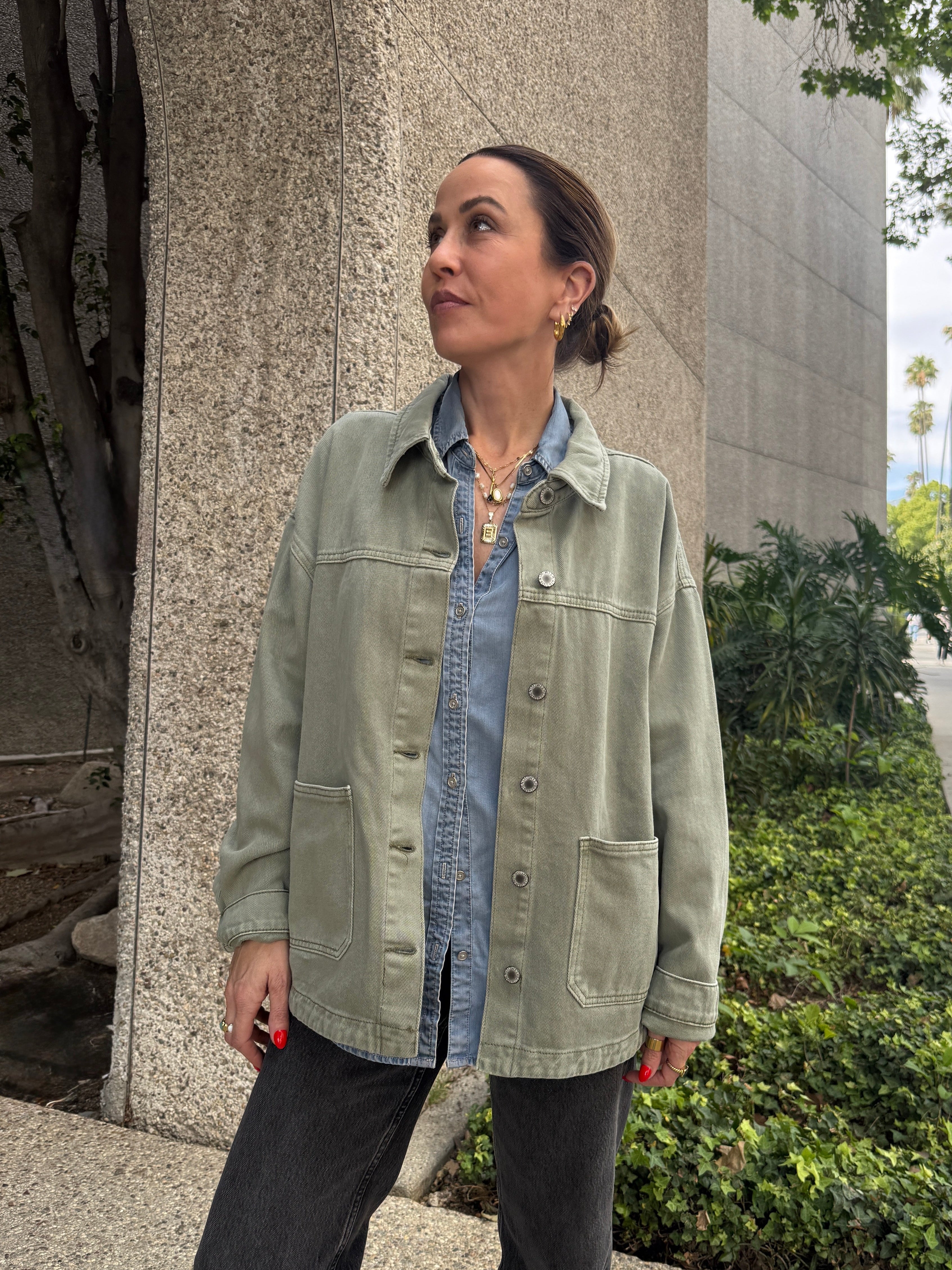Woman standing on a sidewalk wearing a light green jacket over a denim shirt.