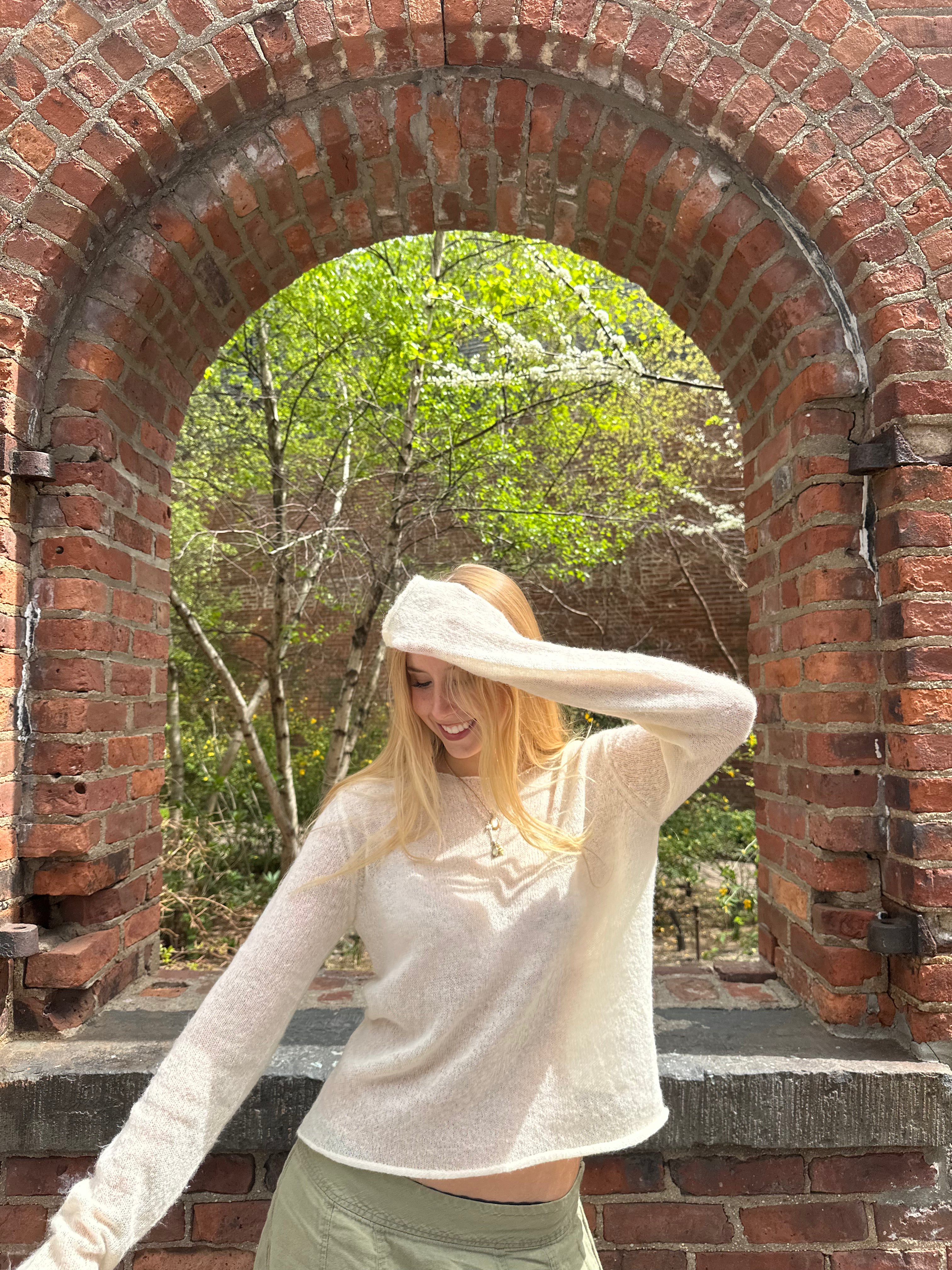 Woman standing under a brick archway with trees in the background wearing cream sweater