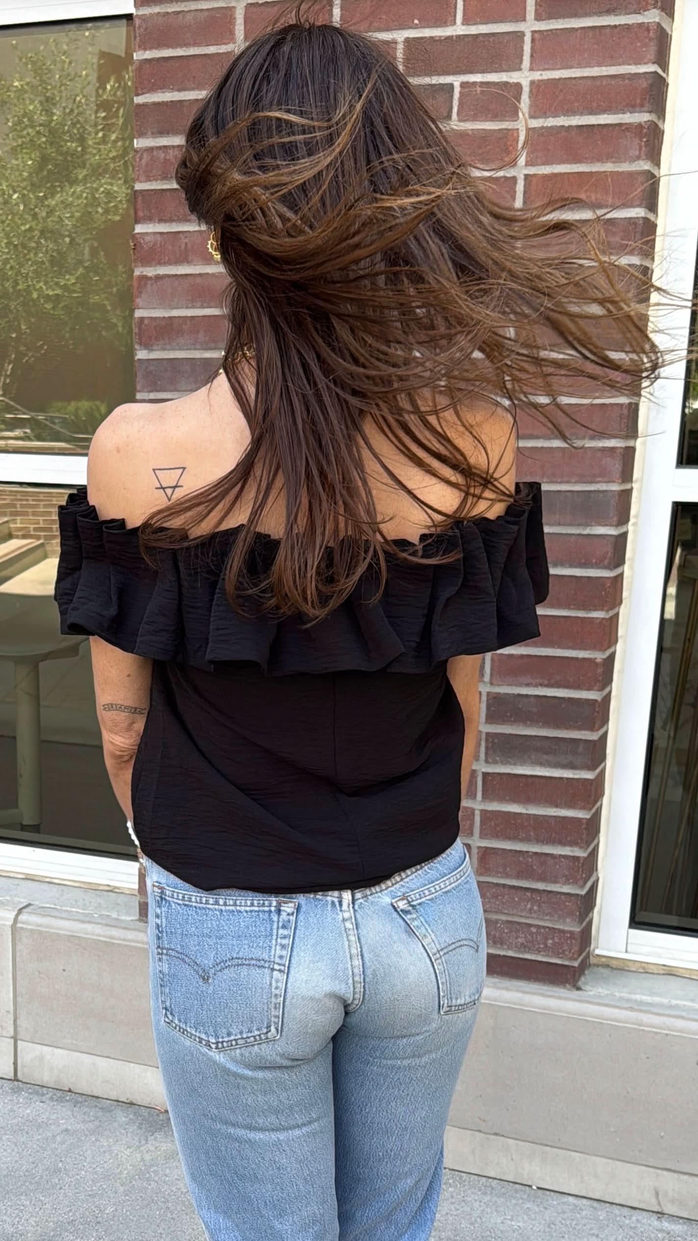 Woman wearing a black off the shoulder ruffle top with blue jeans with pockets behind a brick wall. 