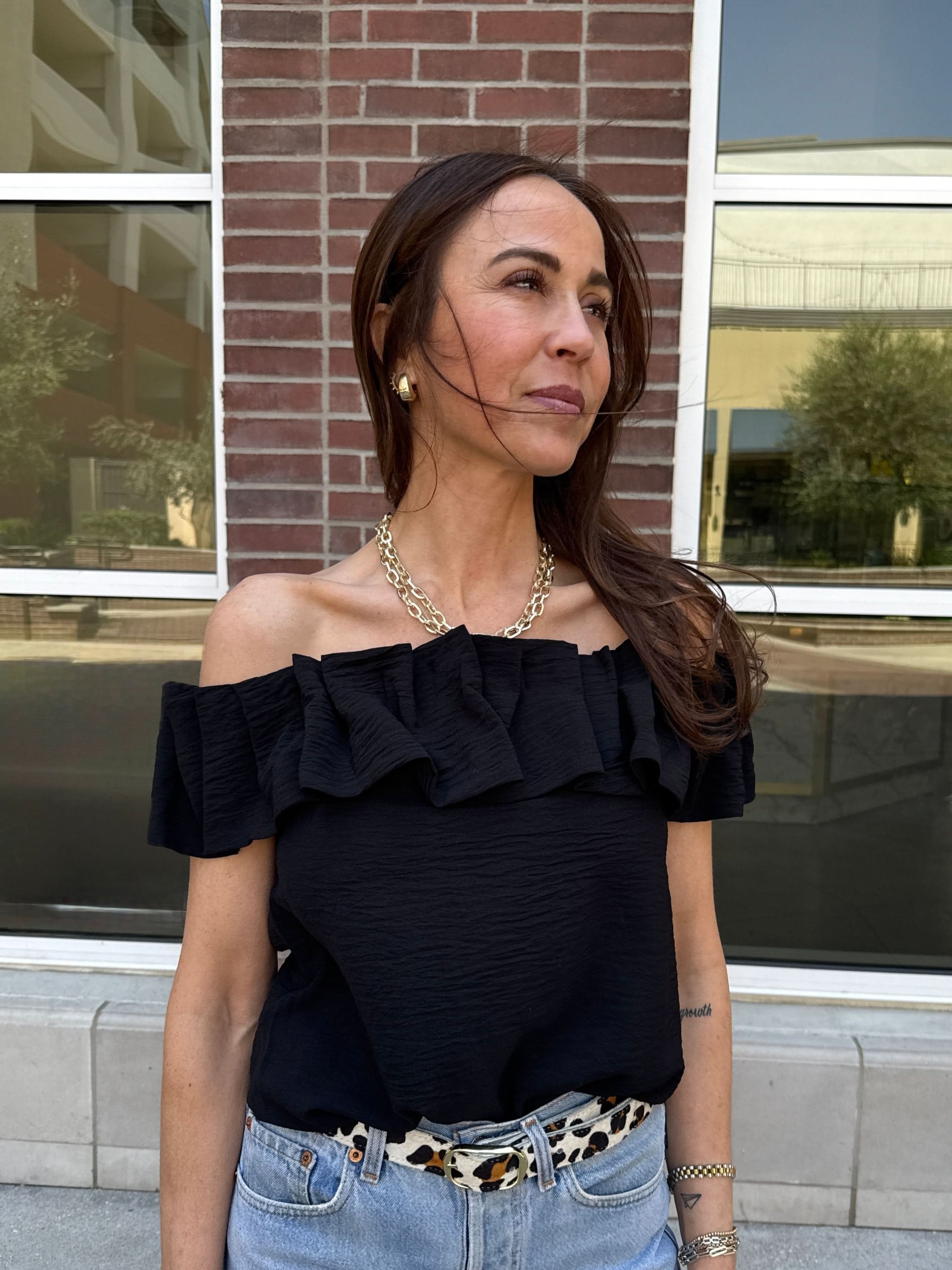 Woman wearing a black off the shoulder ruffle top behind a brick wall. 