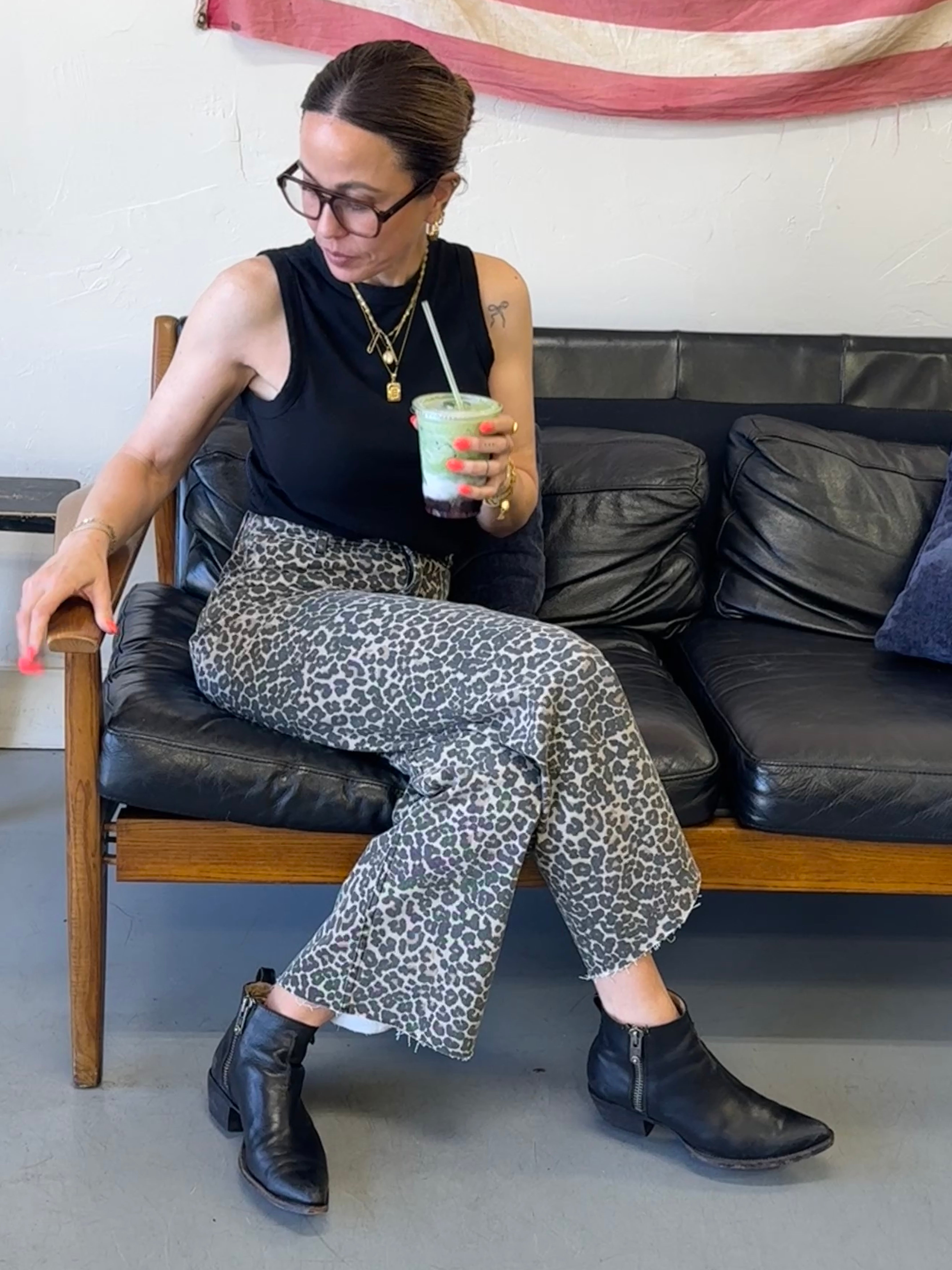 Woman sitting on a black leather couch holding a drink, wearing a black sleeveless top and patterned pants.
