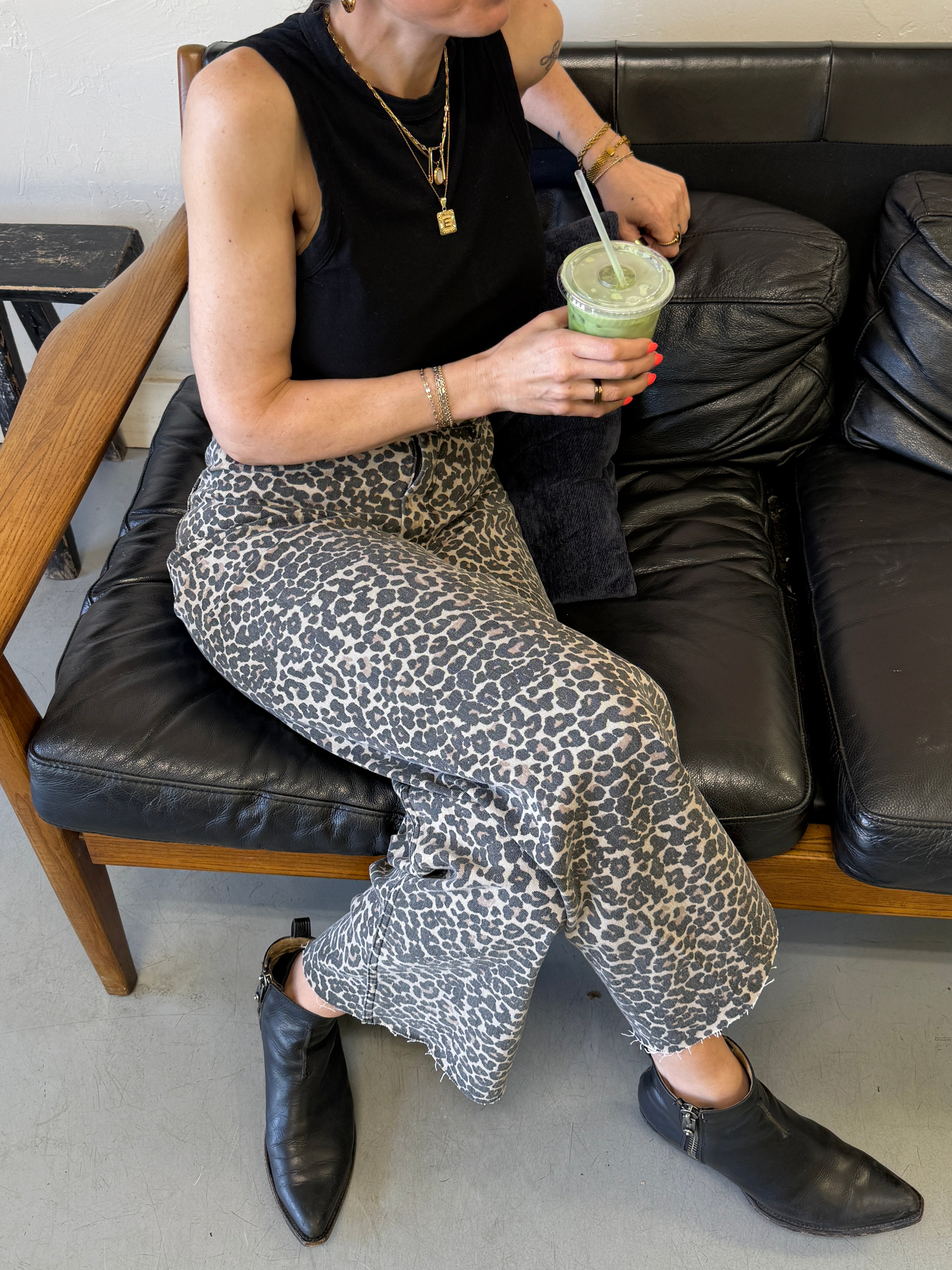 Person sitting on a couch holding a green drink, wearing a black sleeveless top and leopard print pants.