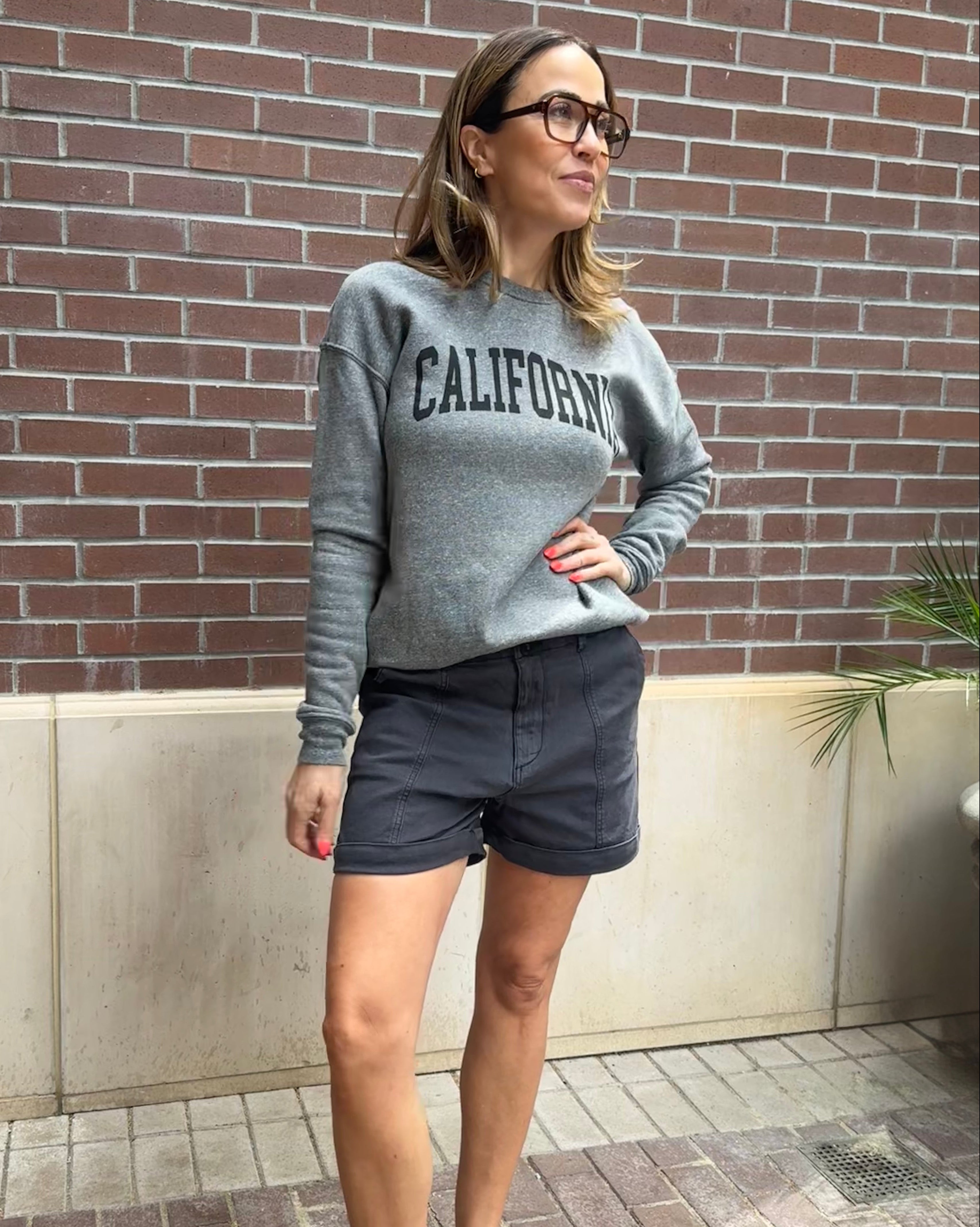 Woman wearing a gray sweatshirt with 'California' text and shorts, standing against a brick wall.
