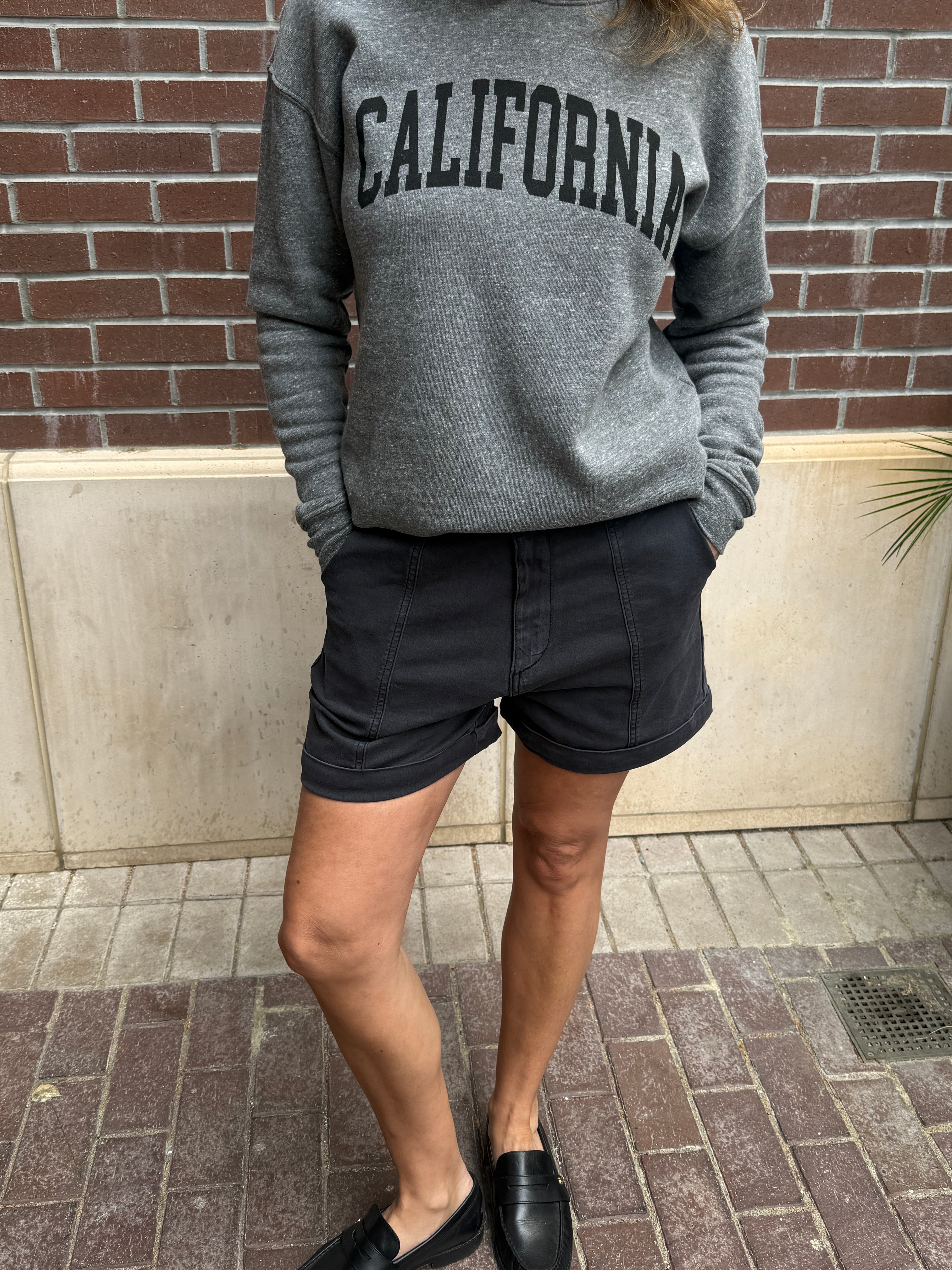 Person wearing a gray sweatshirt with 'California' text and black shorts against a brick wall.