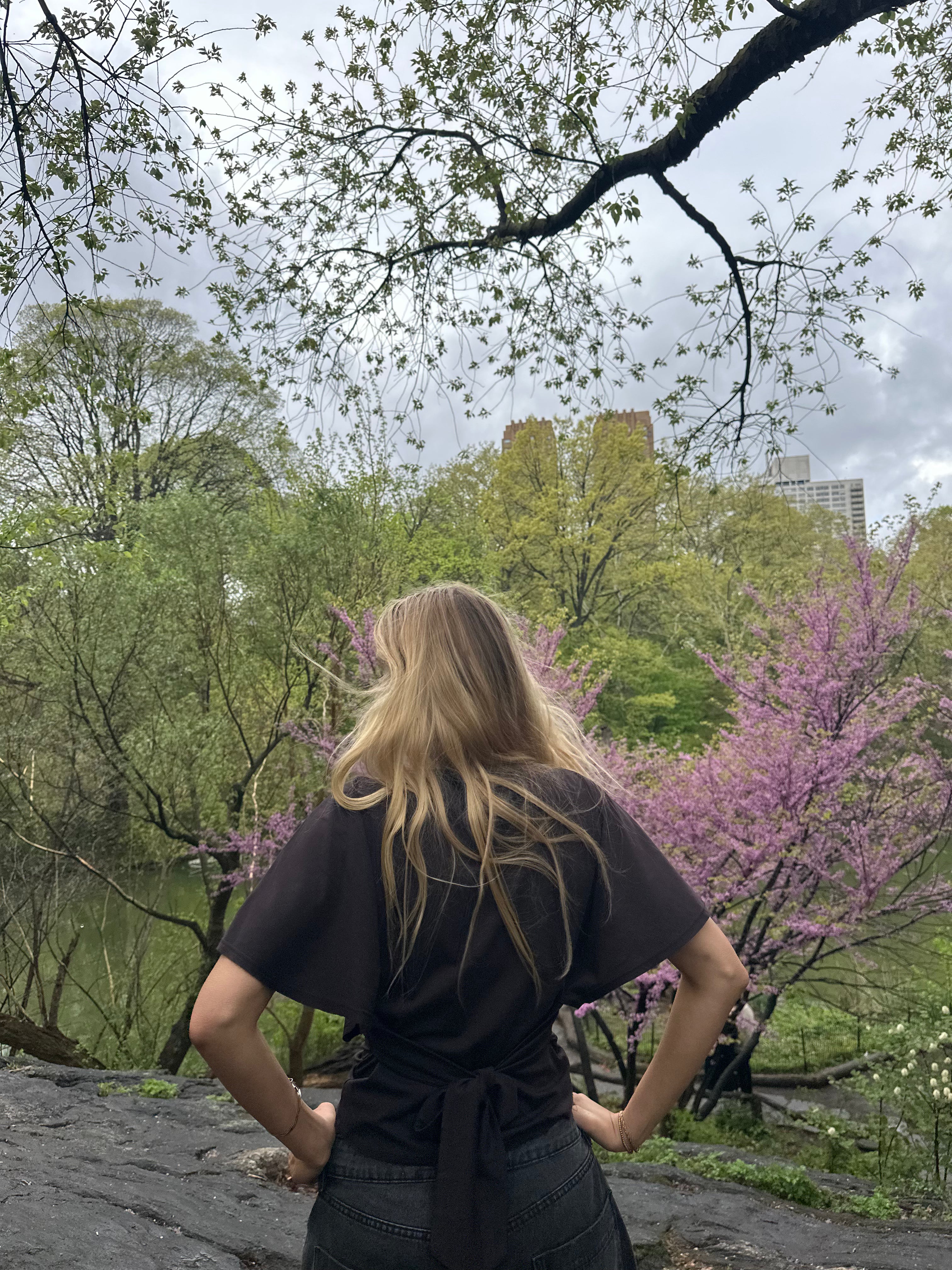 Person with long blonde hair standing in a park with trees and buildings in the background