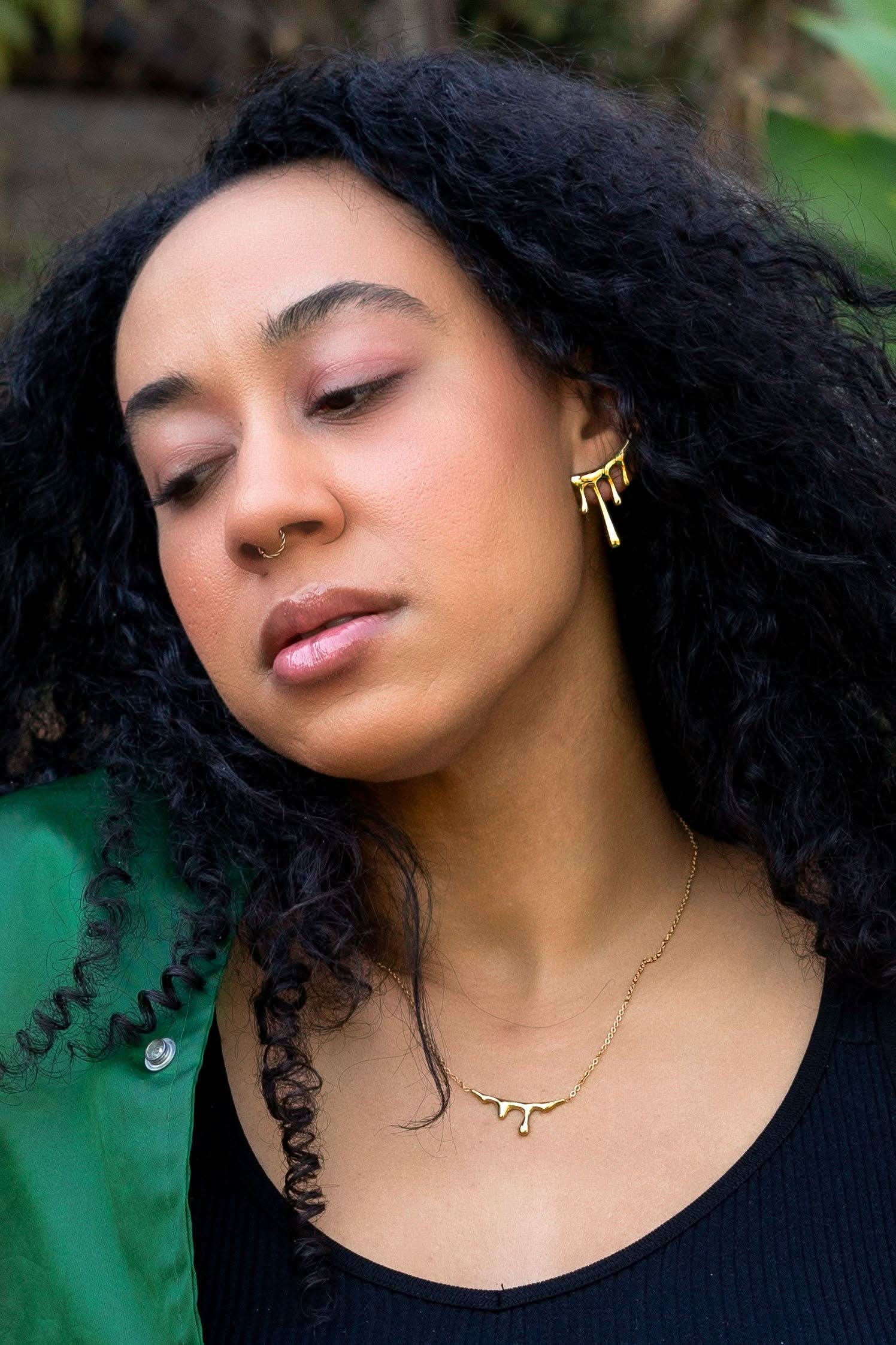 Woman with curly hair wearing gold earrings and a necklace, with a blurred natural background