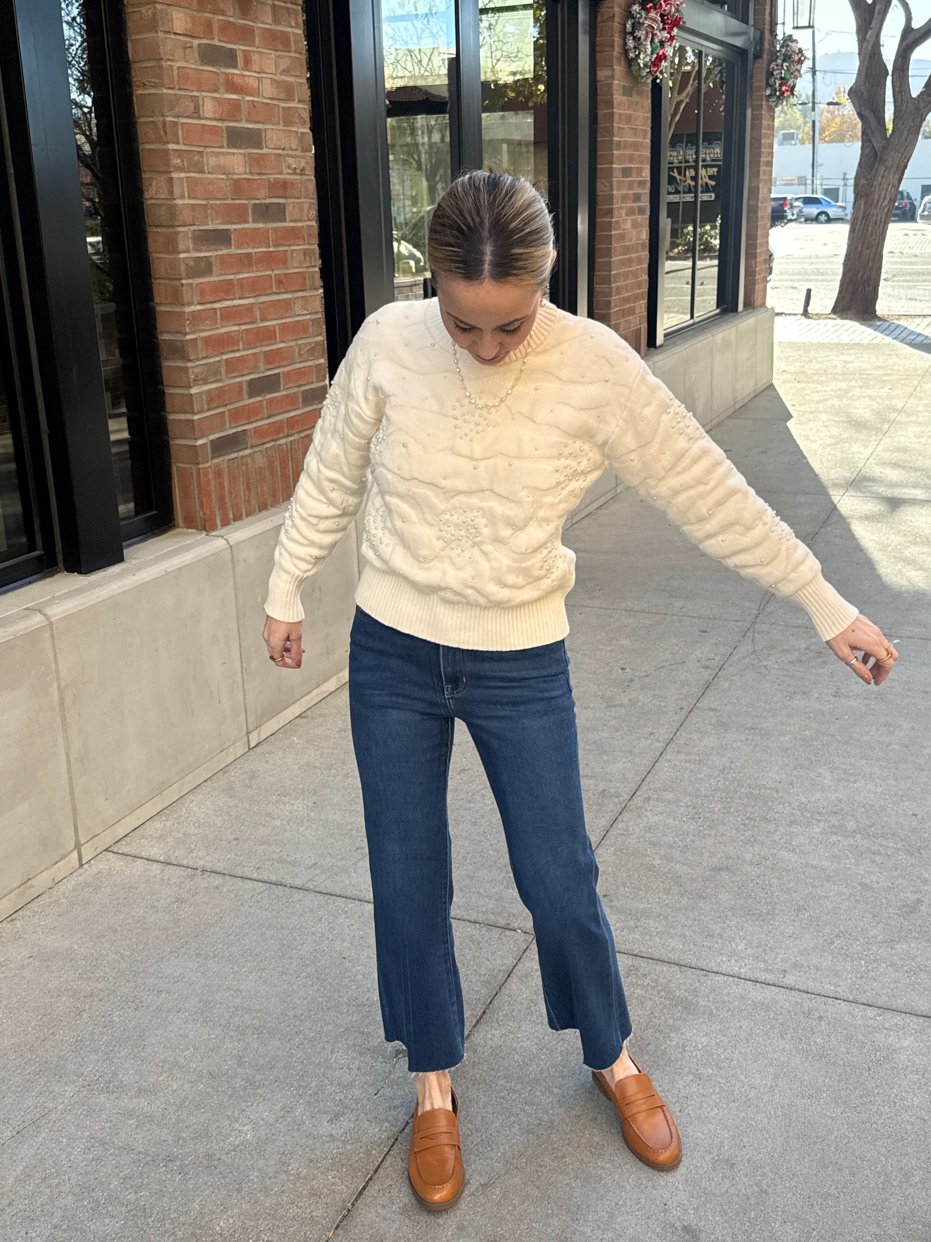 Person wearing a cream sweater, blue jeans, and brown shoes on a sidewalk.