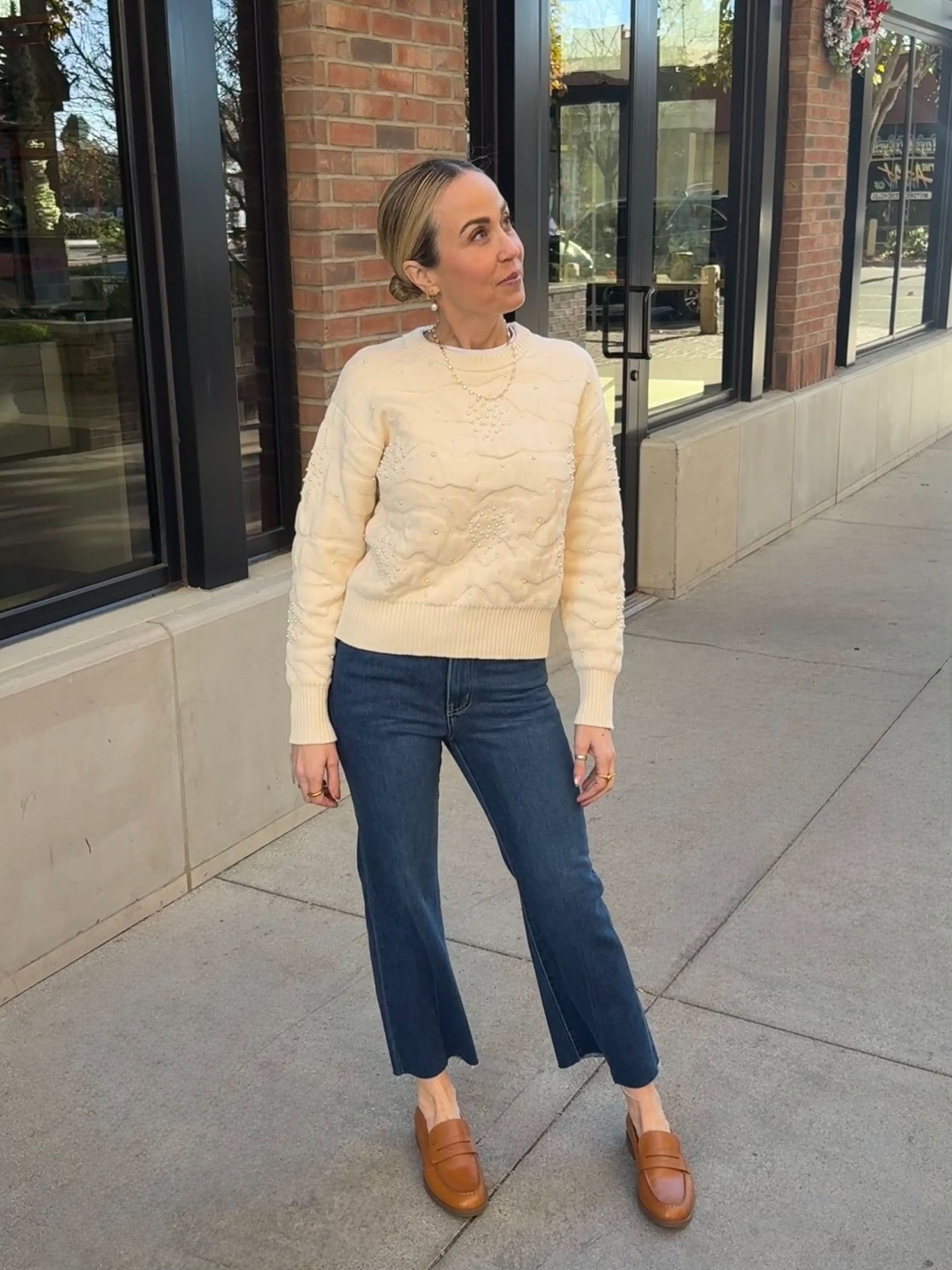 Woman wearing a cream sweater and blue jeans standing on a sidewalk.