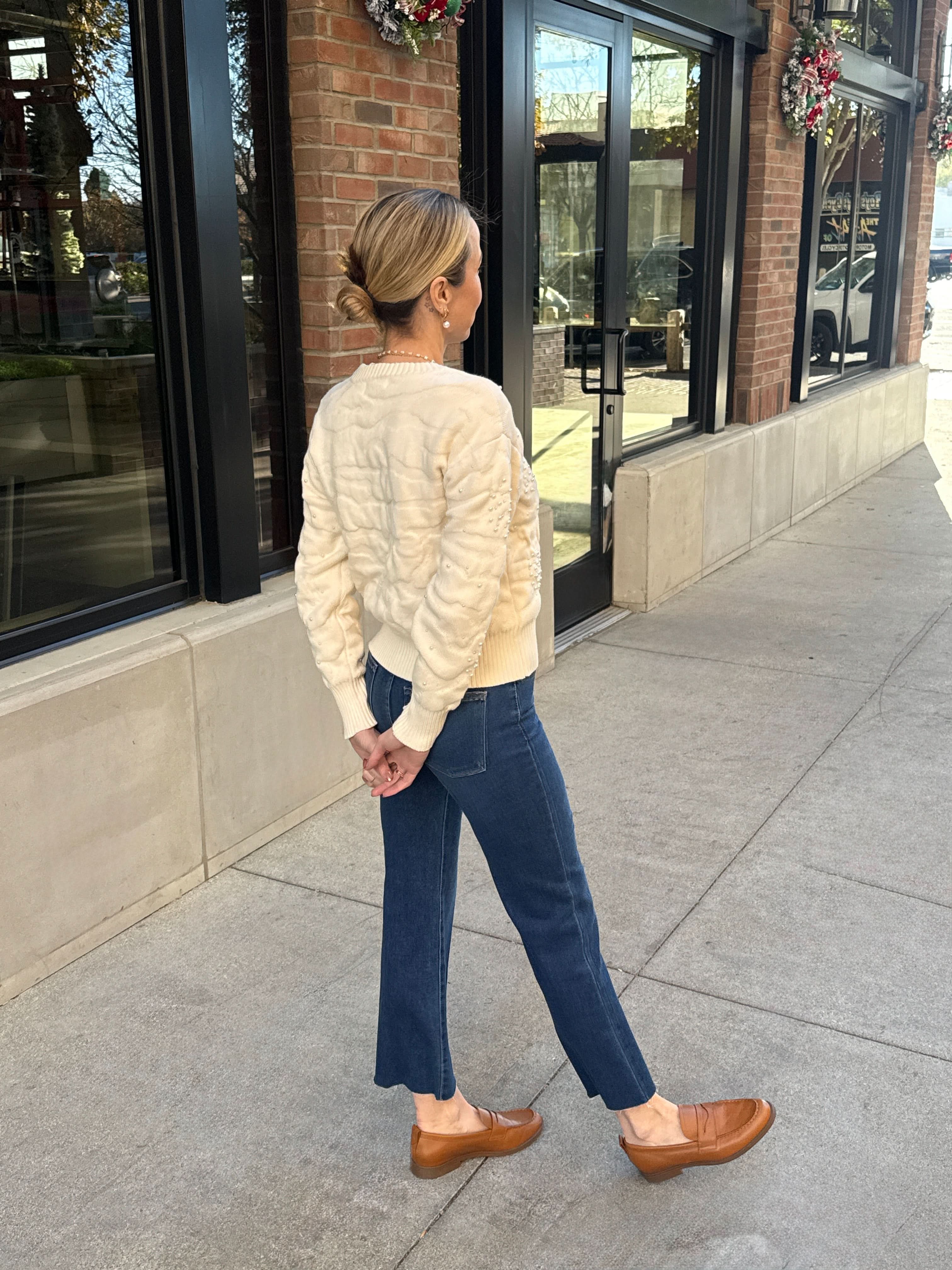 Woman walking on a sidewalk wearing a cream sweater, blue jeans, and brown shoes.