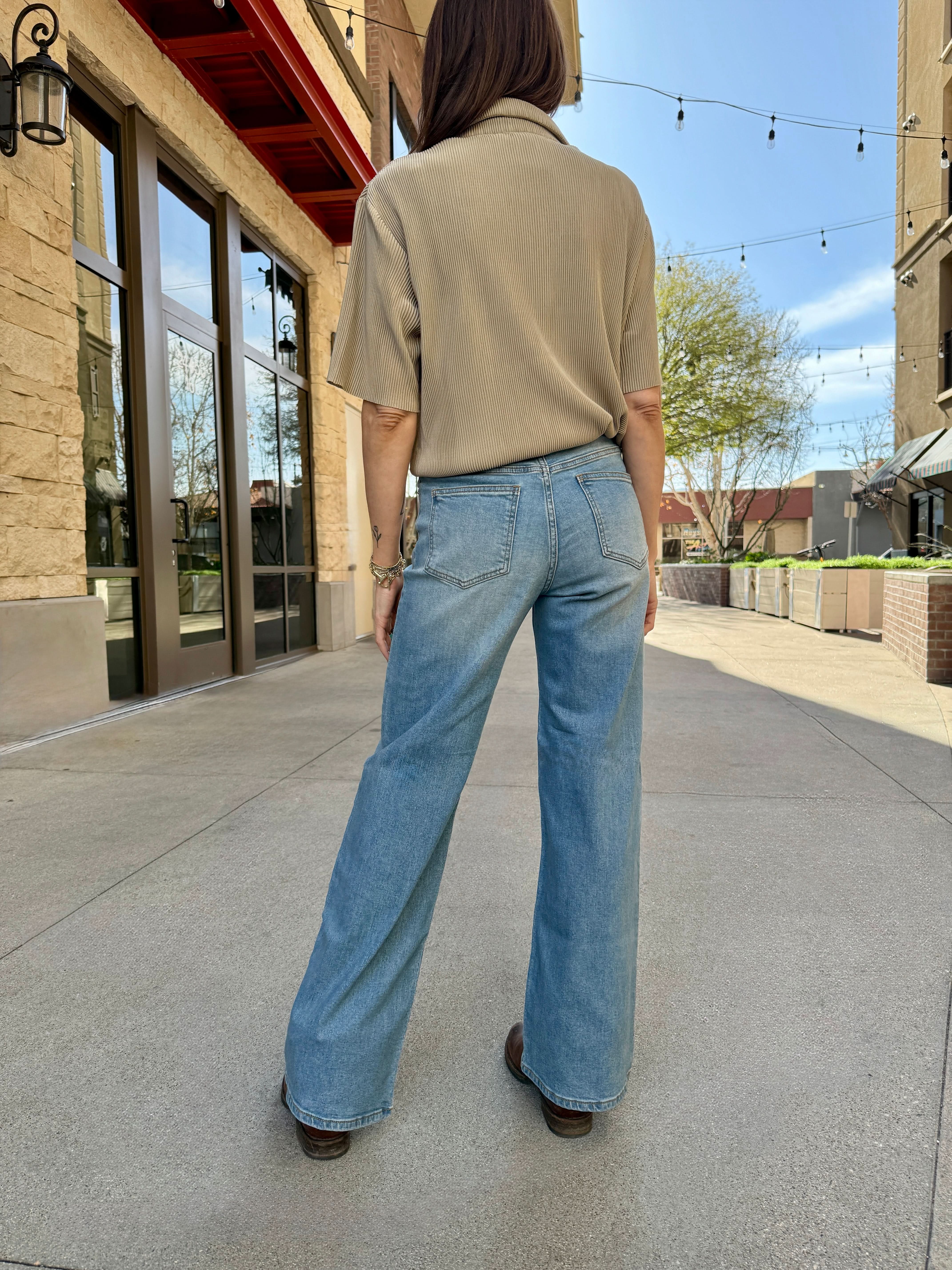 Person wearing a beige shirt and blue jeans standing on a sidewalk with buildings in the background.