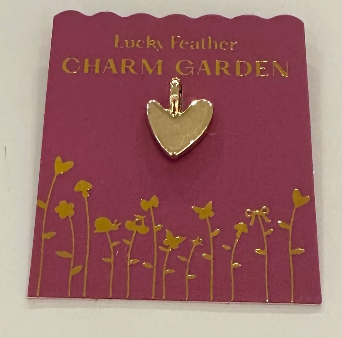 Gold heart-shaped charm on a 'Lucky Feather Charm Garden' card with floral design.
