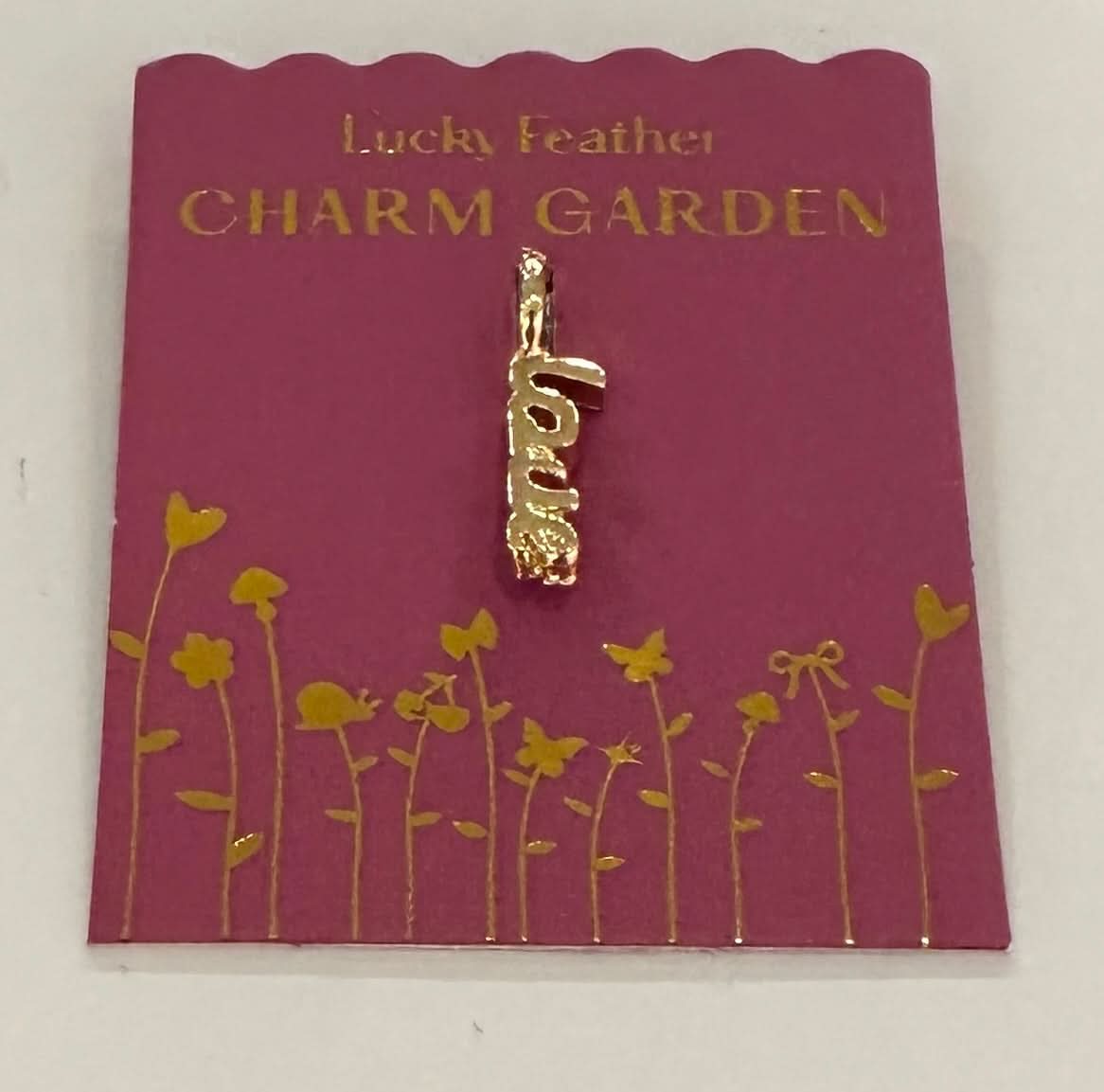 Gold 'love' charm on a 'Lucky Feather Charm Garden' card with floral designs.