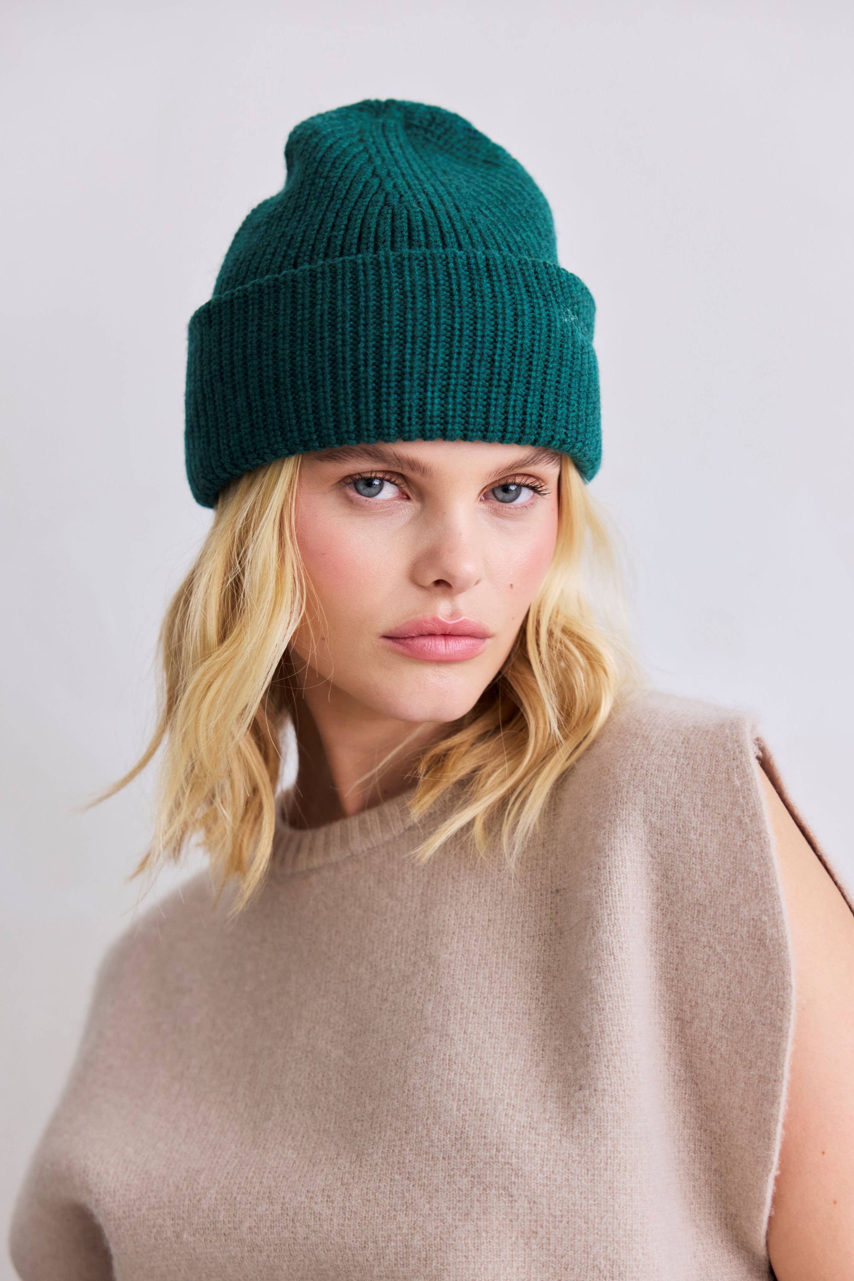 Woman wearing a teal beanie and beige sleeveless top against a white background
